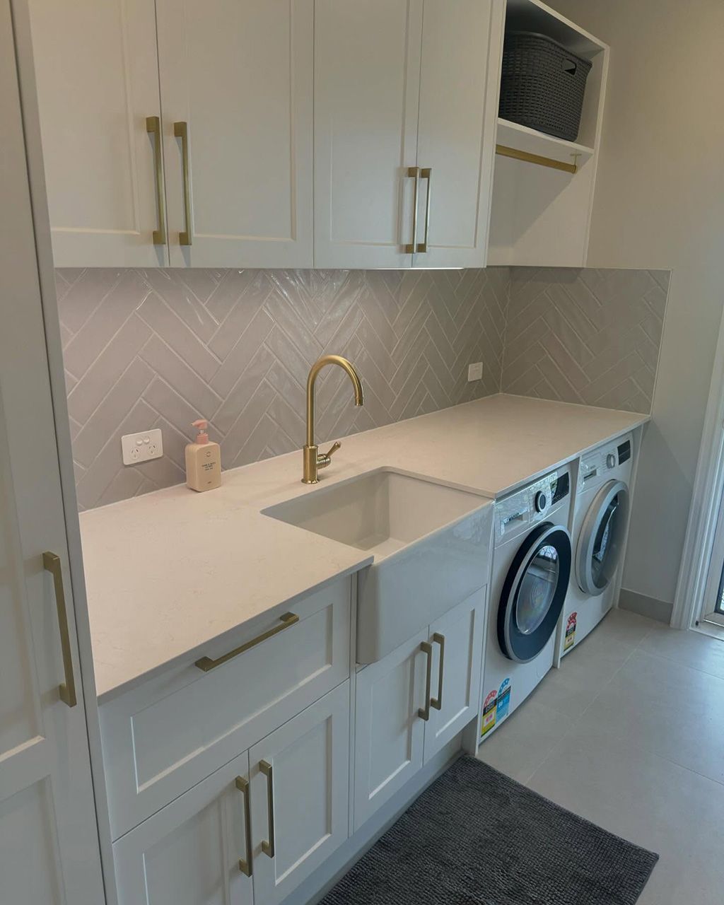 Laundry with stone benchtops on the Gold Coast featuring an undermount sink, brass gooseneck tap and herringbone tile splashback in white shaker cabinetry