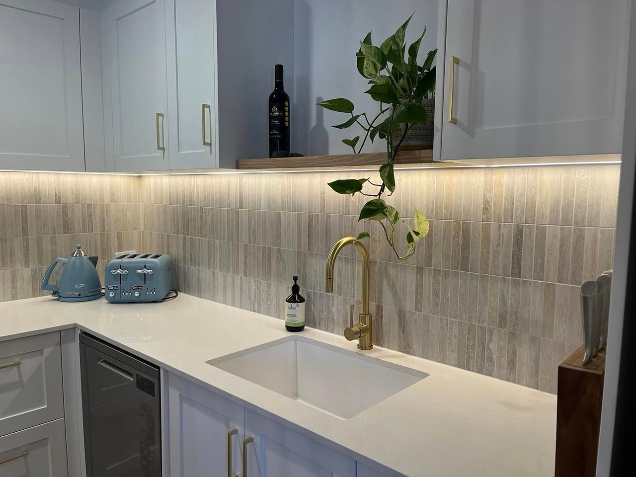 White stone benchtop on the Gold Coast with an undermount sink, brushed brass mixer tap and neutral tile splashback in a light blue shaker kitchen