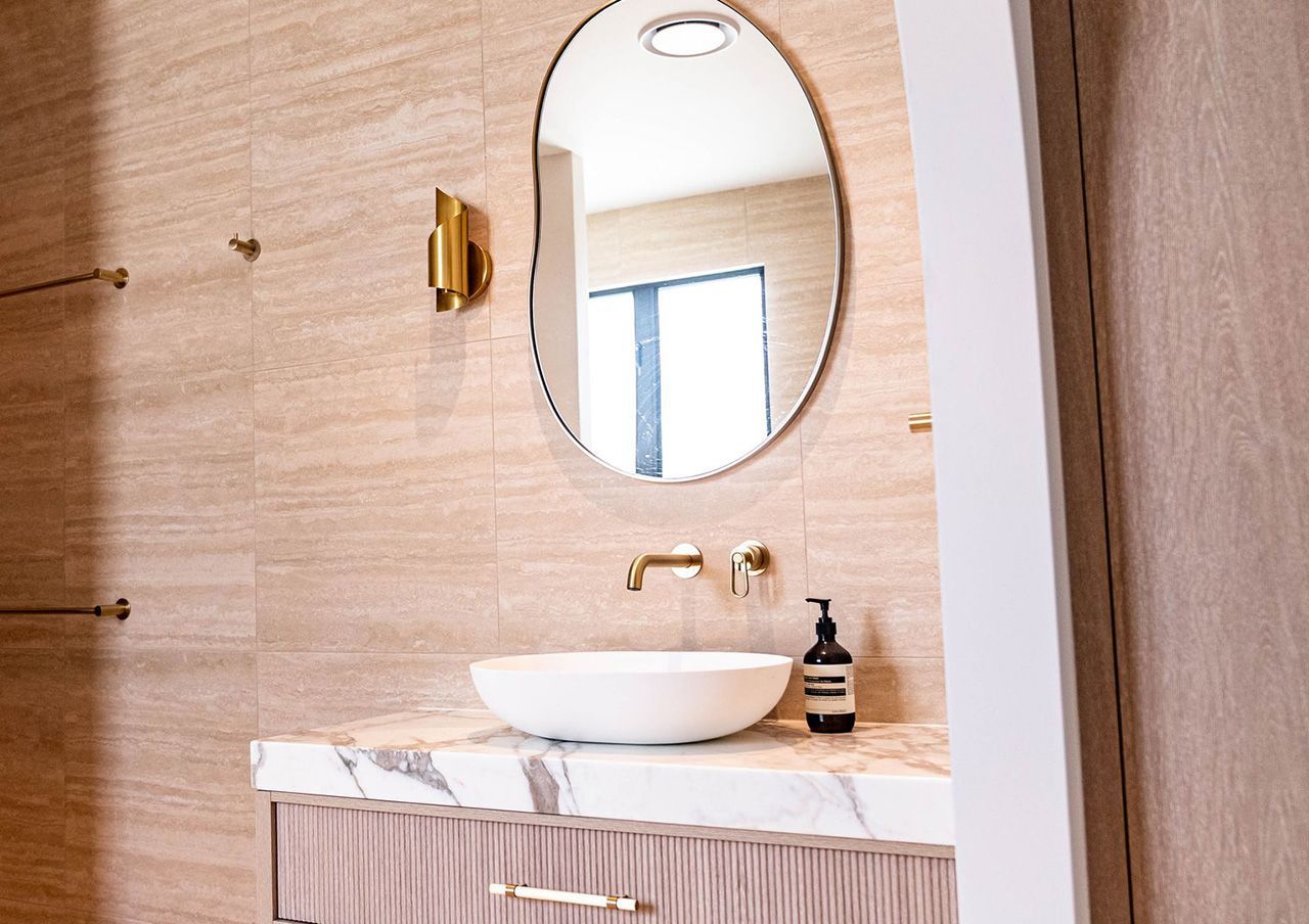 Marble stone benchtop on a Gold Coast bathroom vanity with a vessel basin, brass wall-mounted tap and fluted cabinet below an organic-shaped mirror