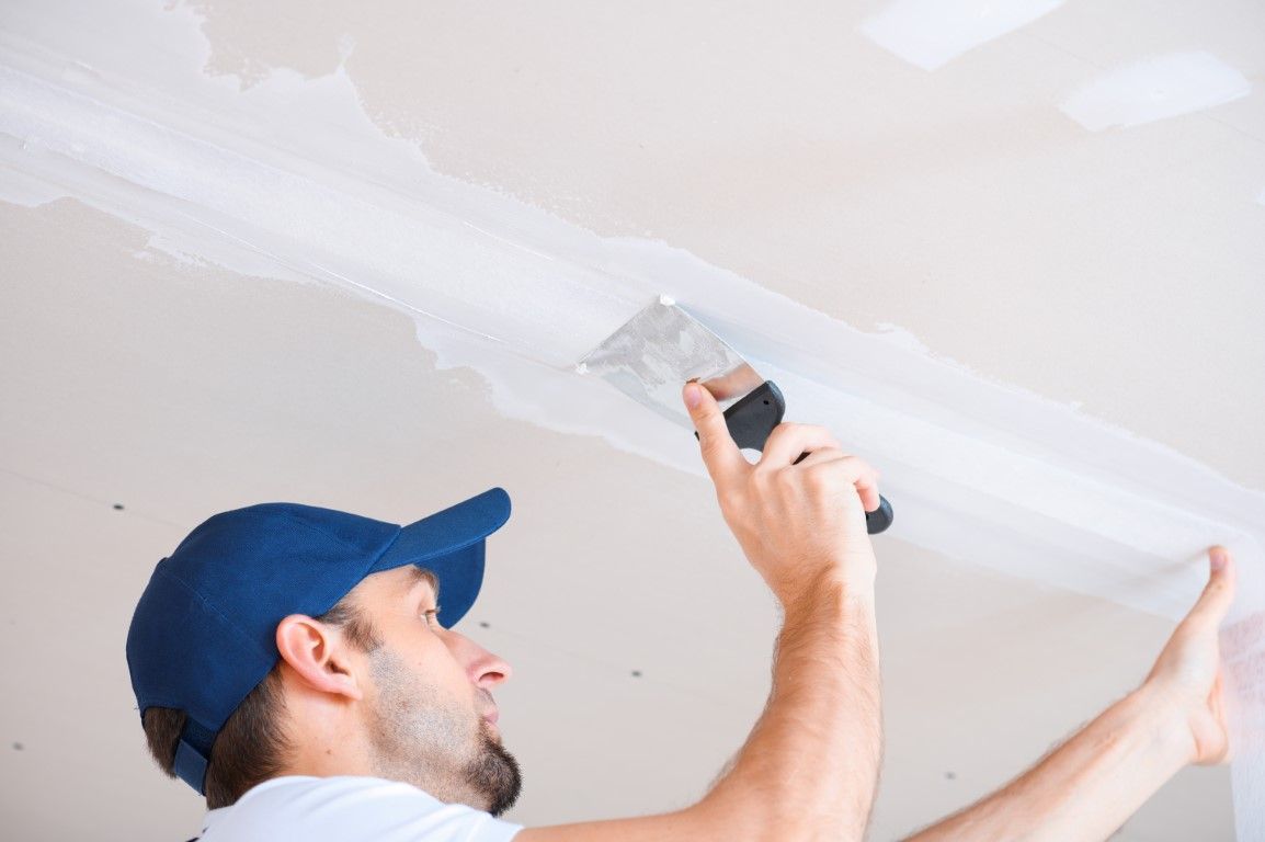 Person using a putty knife to smooth drywall compound on a ceiling while standing on a ladder