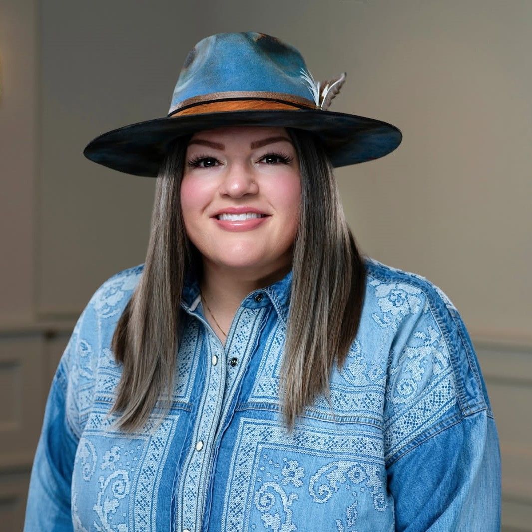 Woman in a blue patterned shirt and hat, smiling against a neutral indoor background