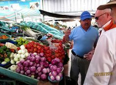 Galapagos Two men are standing in front of a vegetable stand at a market.
