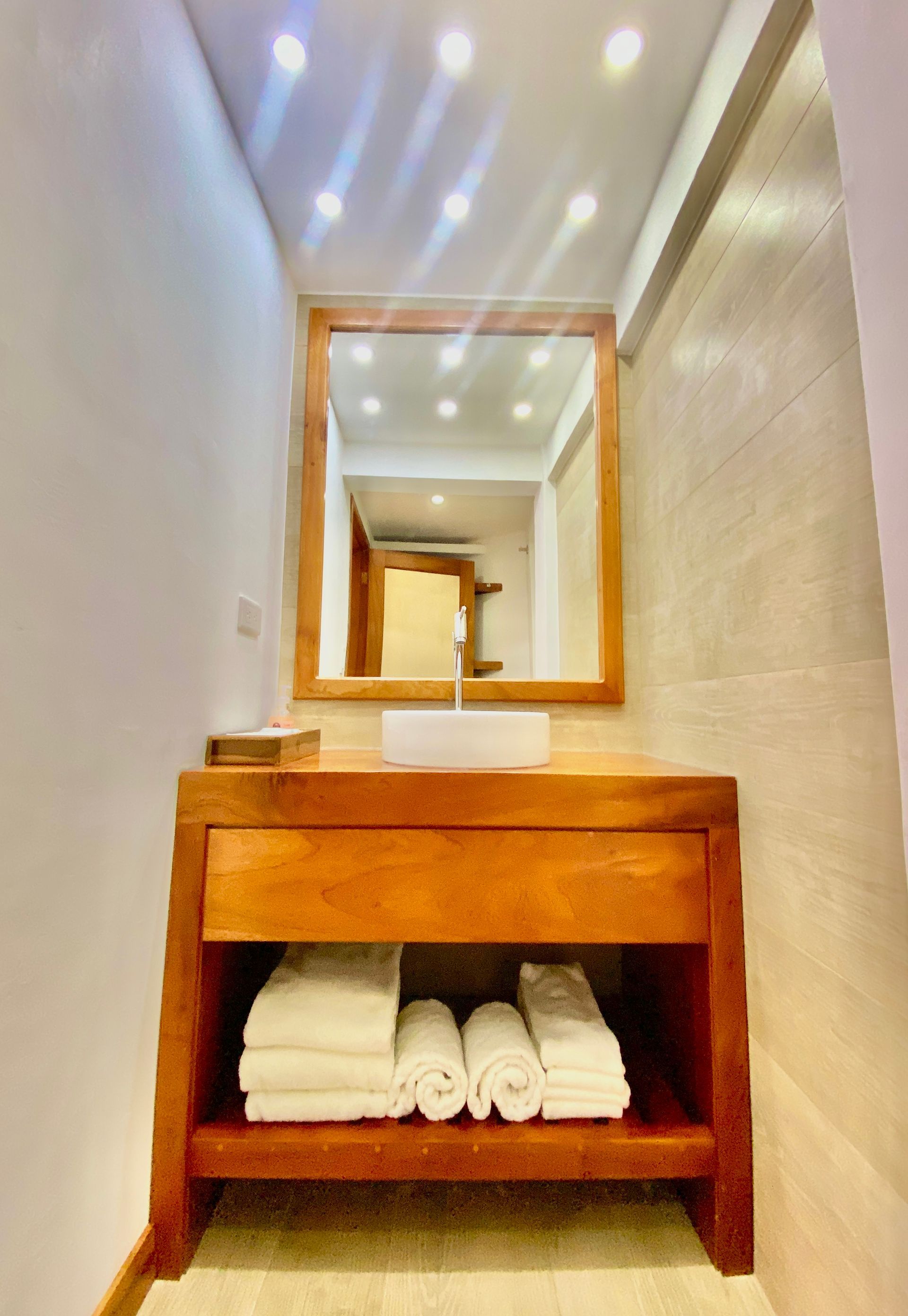A Galapagos hotel wooden vanity with a mirror, a white sink, and rolled white towels sits in a narrow bathroom.