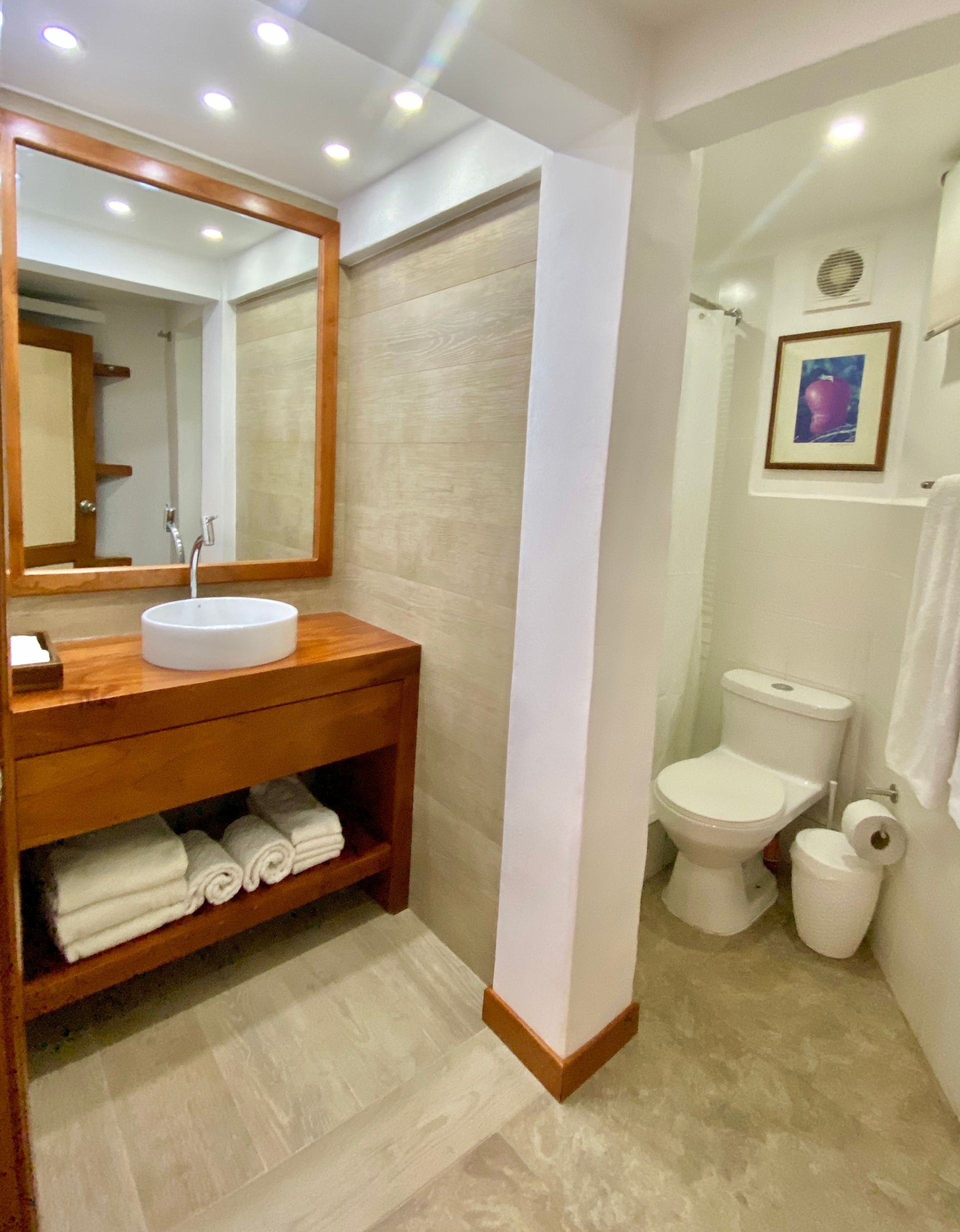 Hotel bathroom with wooden vanity, round sink, large mirror, and toilet in a white tiled alcove.