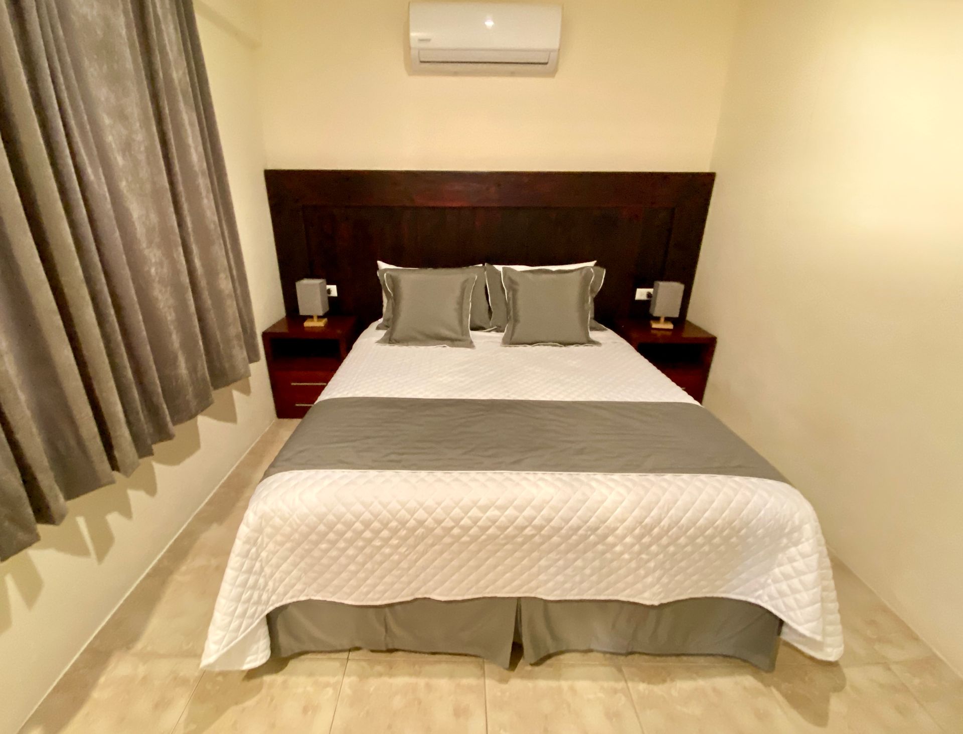 A Galapagos hotel bedroom with a bed, two nightstands, and an air conditioning unit. The bed has gray and white bedding.
