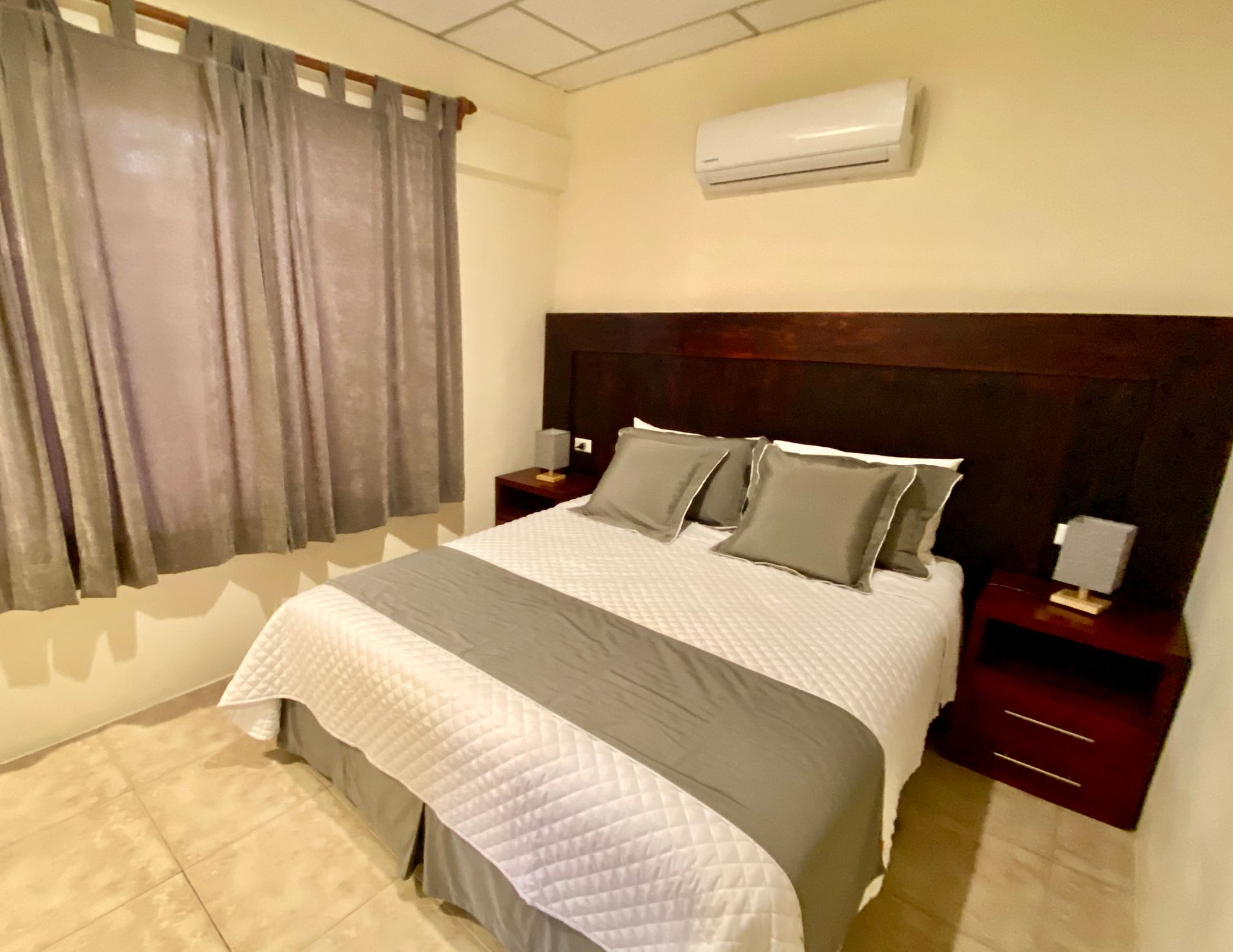 A Galapagos hotel Bedroom with a bed, nightstands, window with gray curtains, and air conditioning unit.