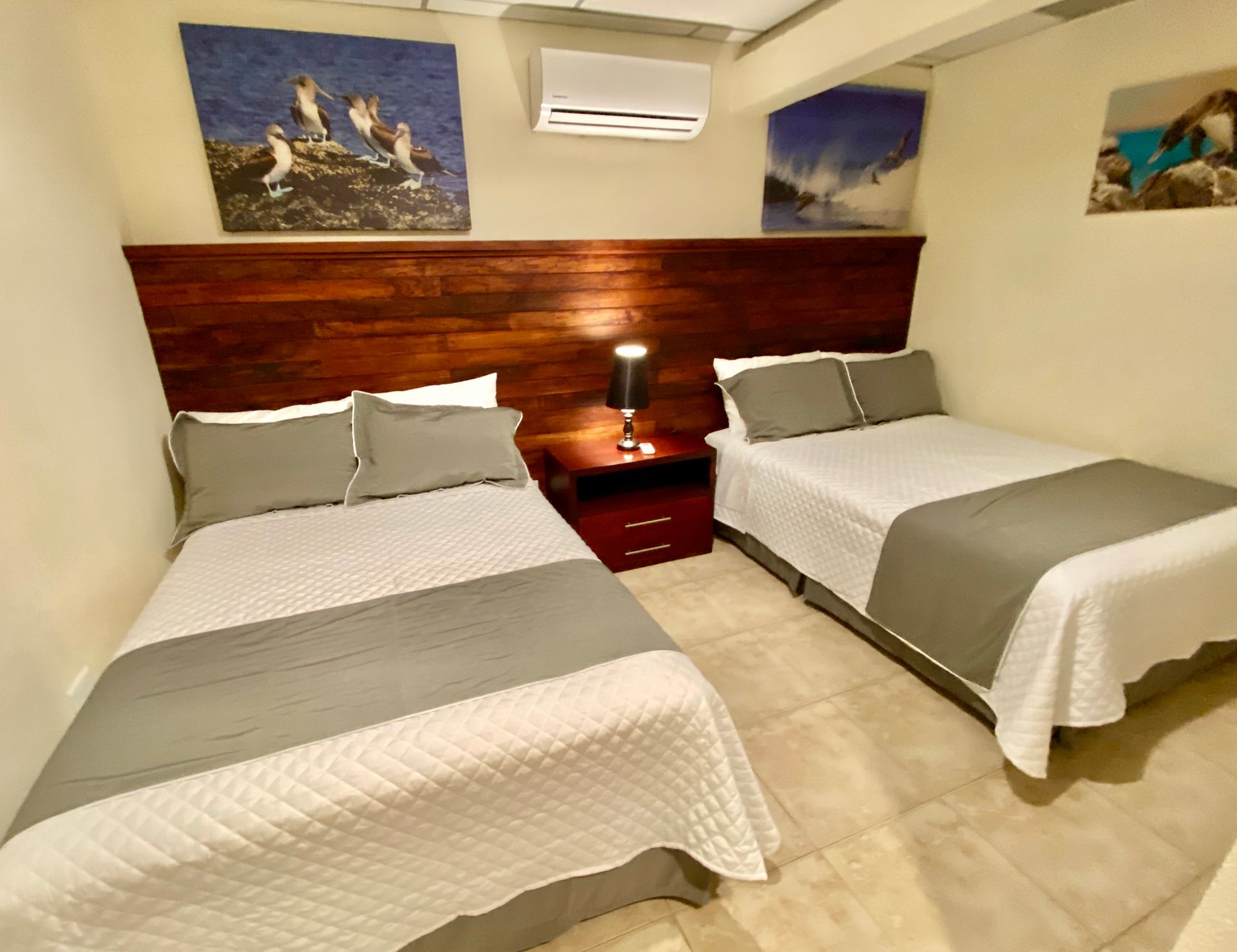 Two double beds in a beige hotel room with a wood headboard, bedside lamp, and wall art