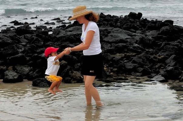Mother and child holding hands in shallow water by rocky shore, wearing hats on a beach