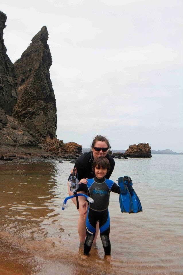 Adult helping child in blue wetsuit stand in shallow water beside rocky beach and sea