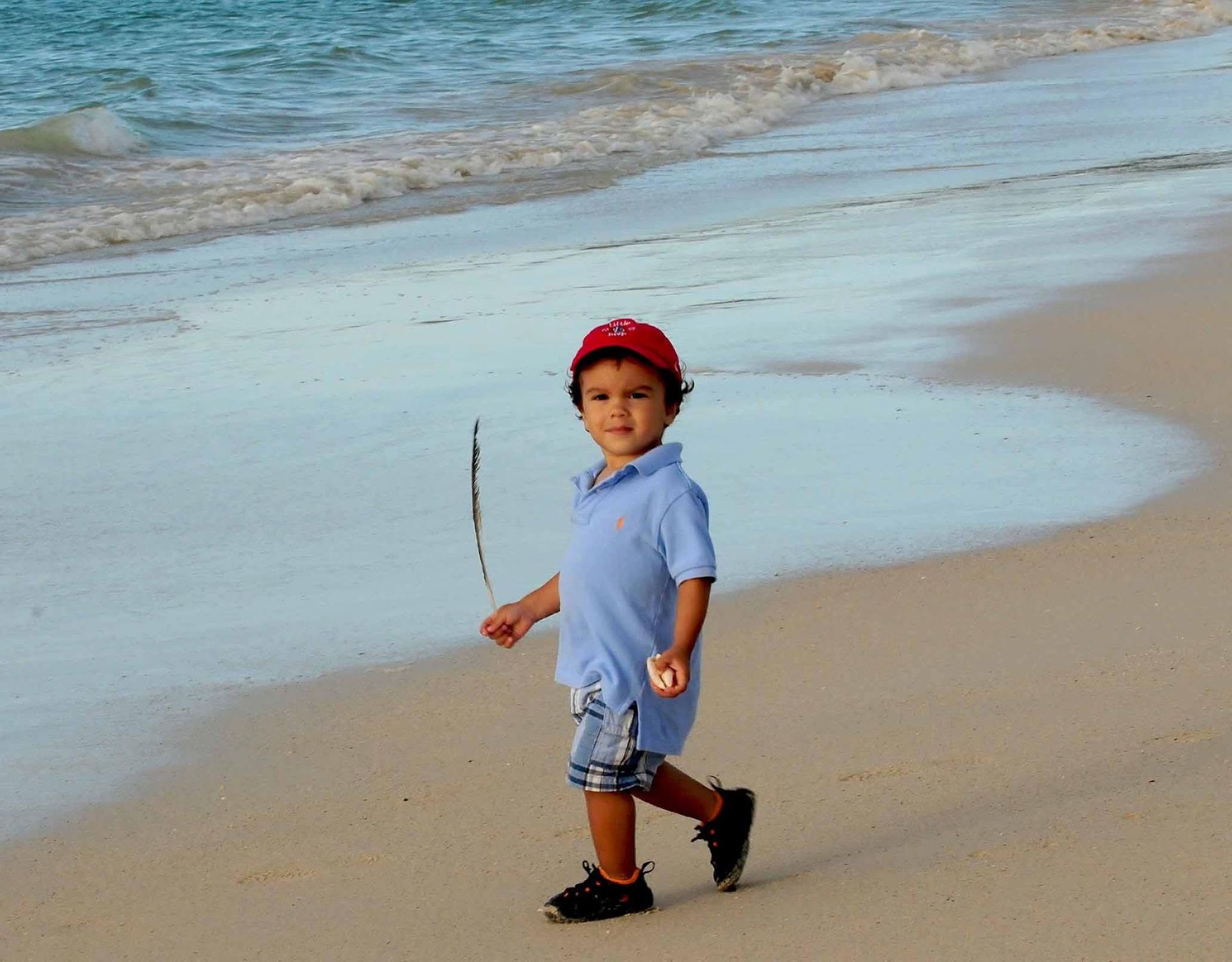 Child in a red cap and blue shirt walking on a beach near the waterline.