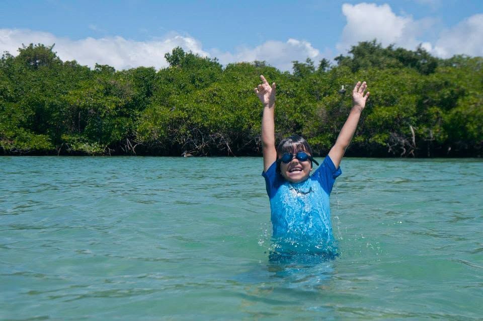 Person in blue shirt raising arms in shallow water near a mangrove-lined shore
