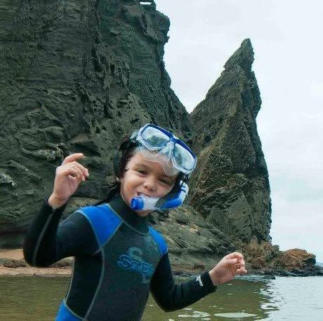 Child in a blue wetsuit and snorkel stands by Pennacle Rock and water.