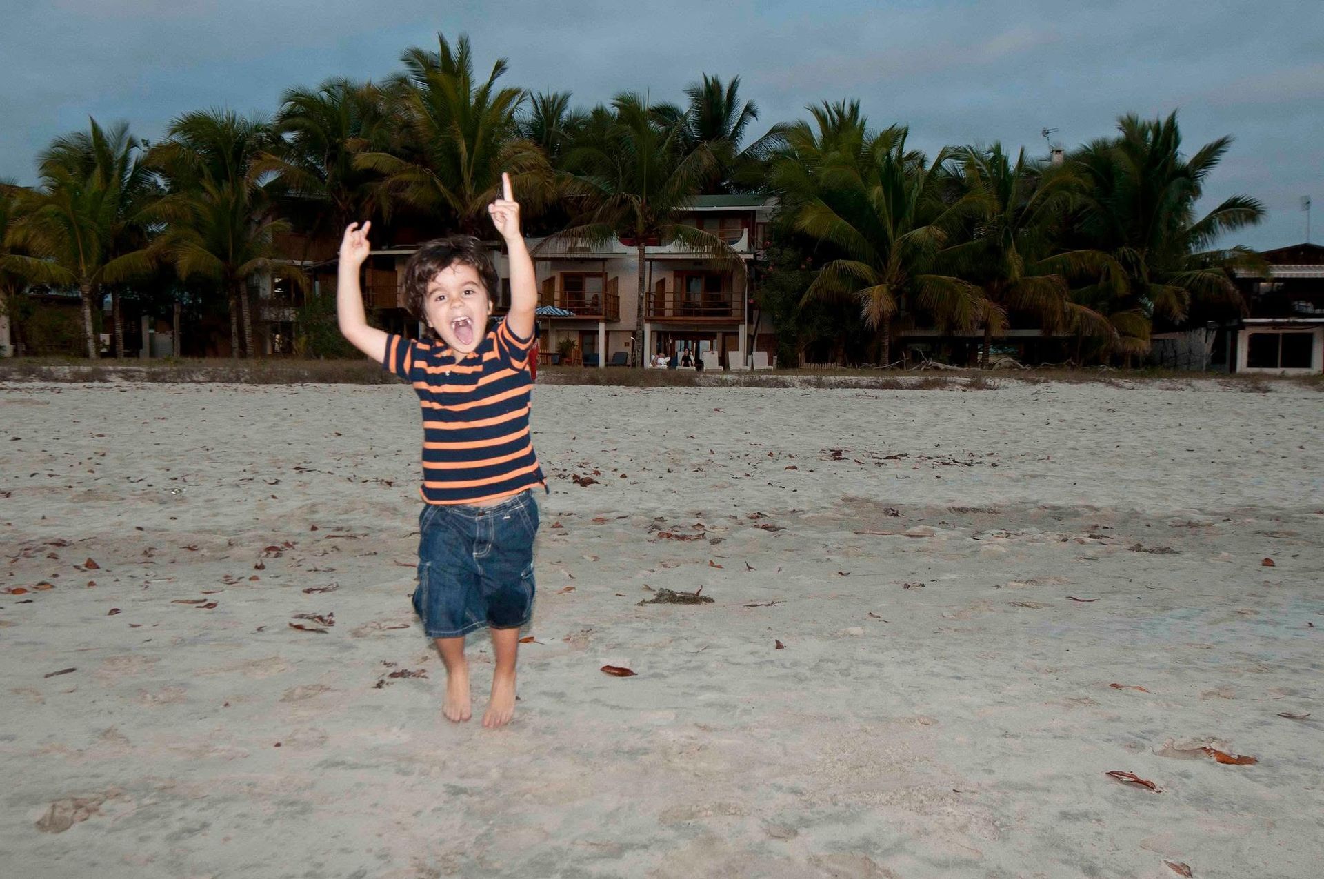 Child in a striped shirt and denim shorts stands on a beach with arms raised, palm trees behind.