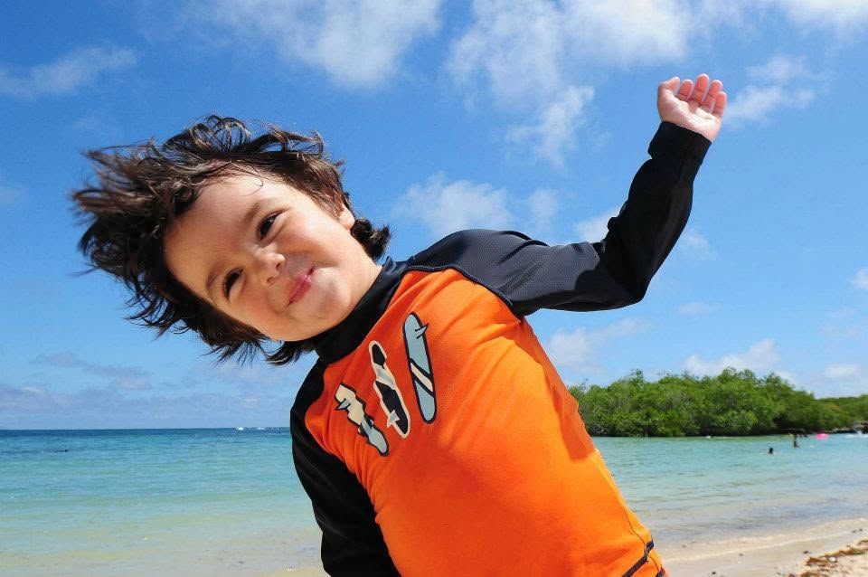 Child in an orange shirt smiling and leaning on a beach with blue sky and ocean in the background