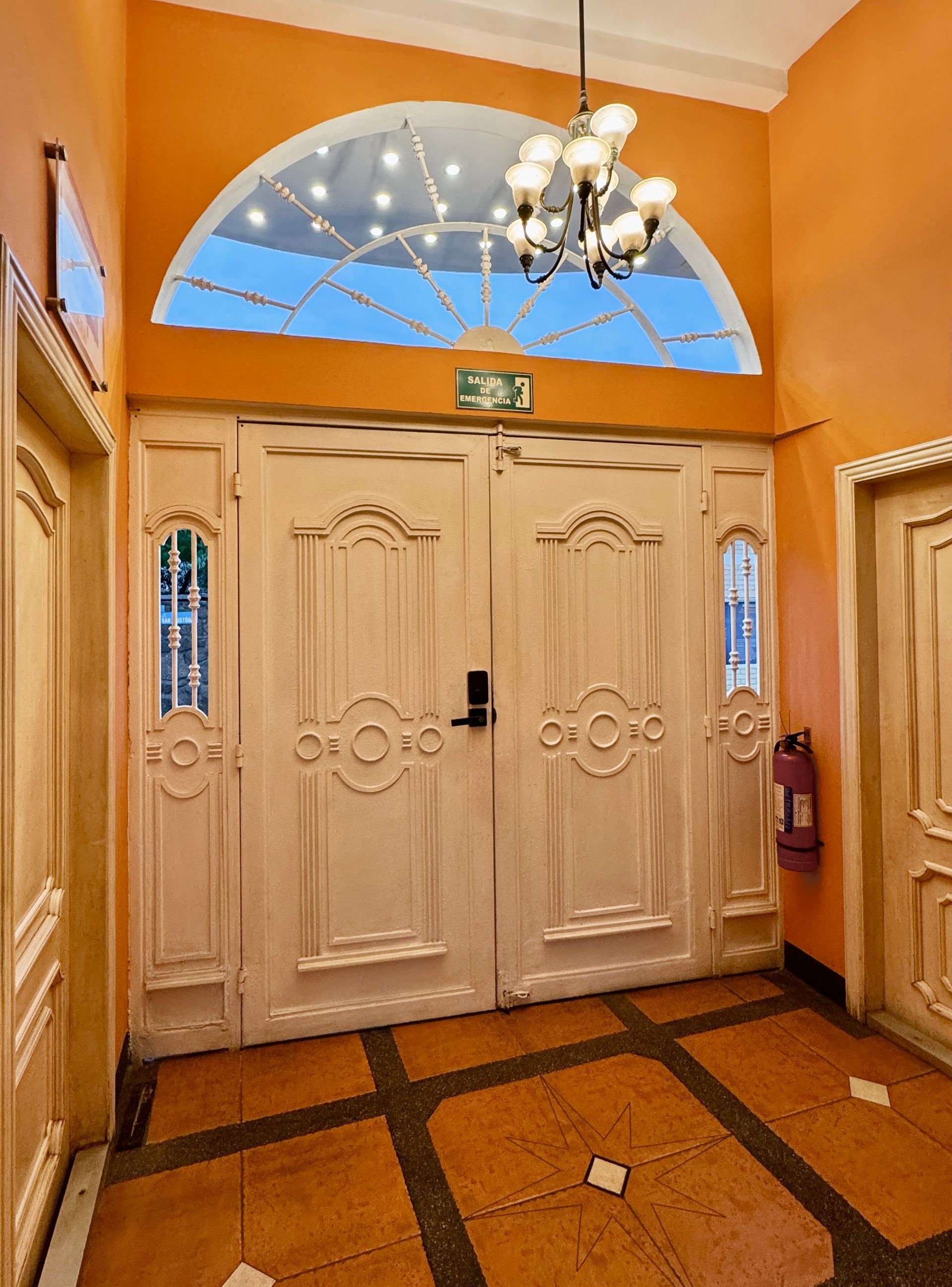 Peach entryway with ornate cream double doors, chandelier, arched window, and tiled floor, Galapagos