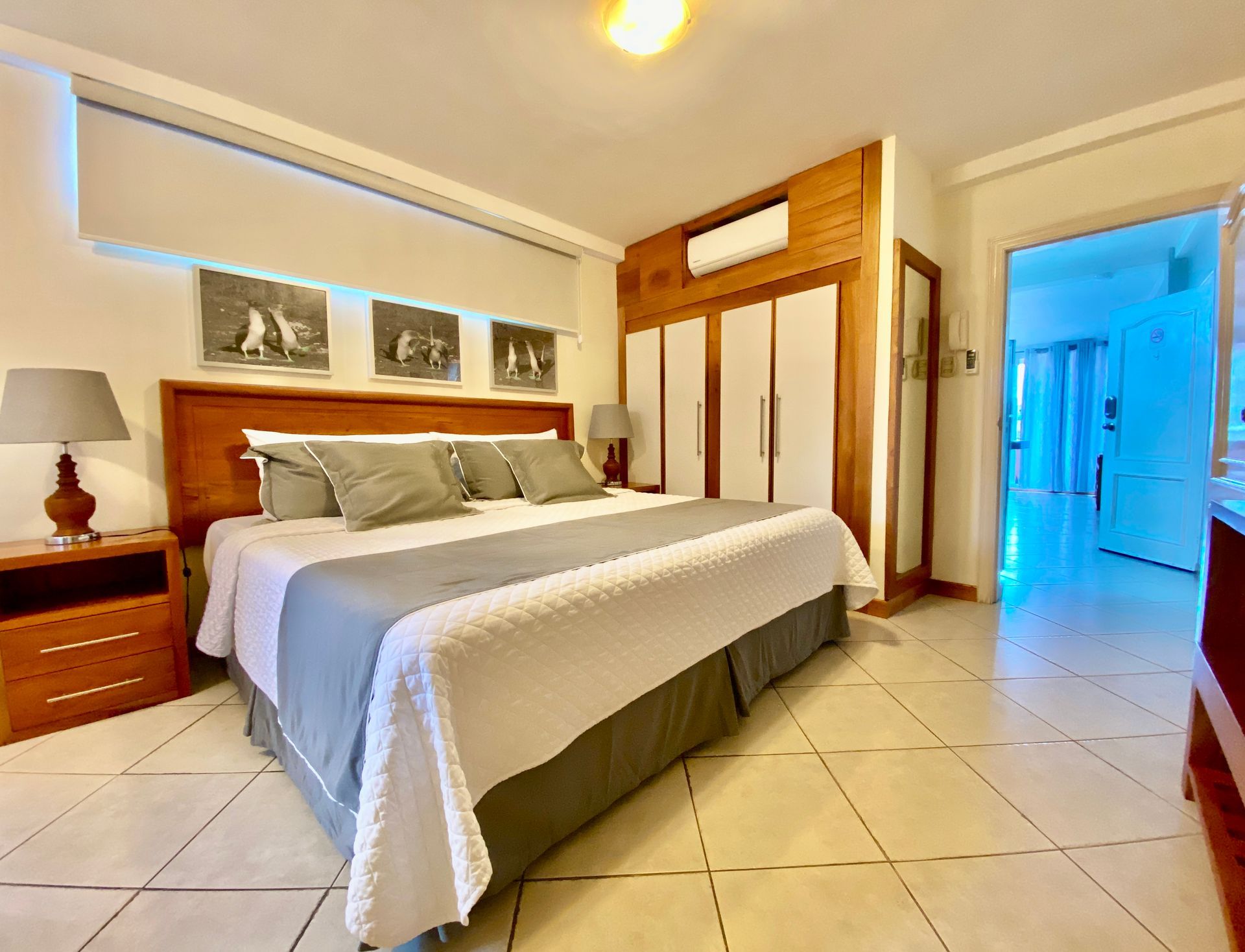 Bright bedroom with a large bed, wooden furniture, tiled floor, and an open doorway to a blue-lit hall