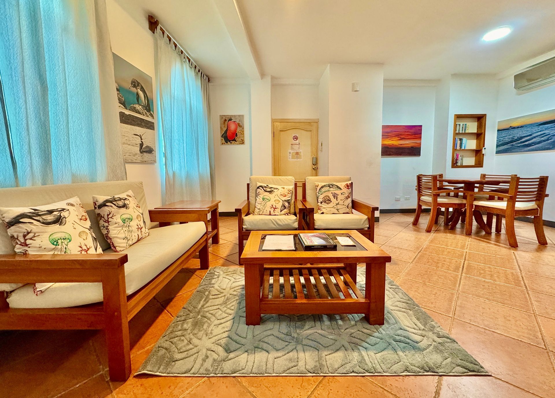 Bright Hotel living-dining room with wooden furniture, blue curtains, and a kitchen counter in the background in the Galapagos Islands.