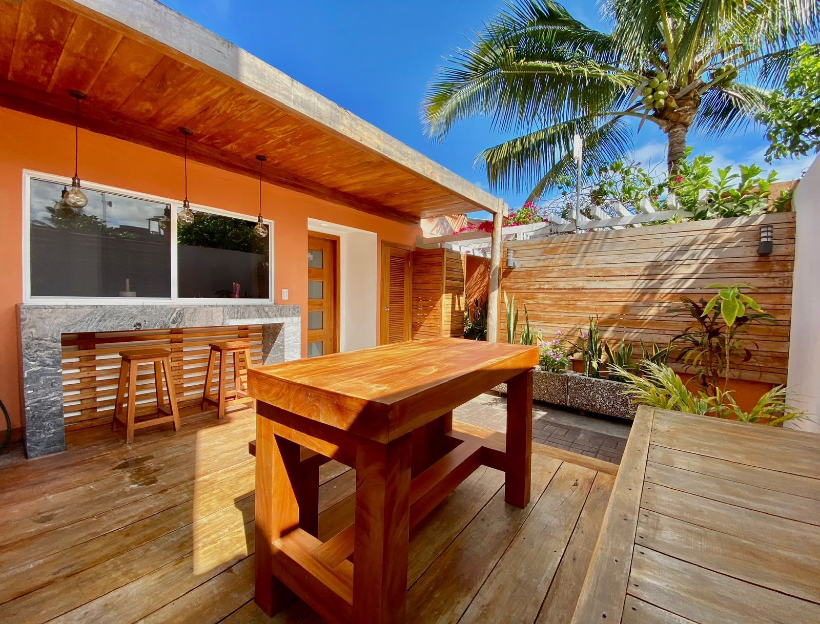 Wooden patio with outdoor table, stools, and palm tree beside a house under bright sun.