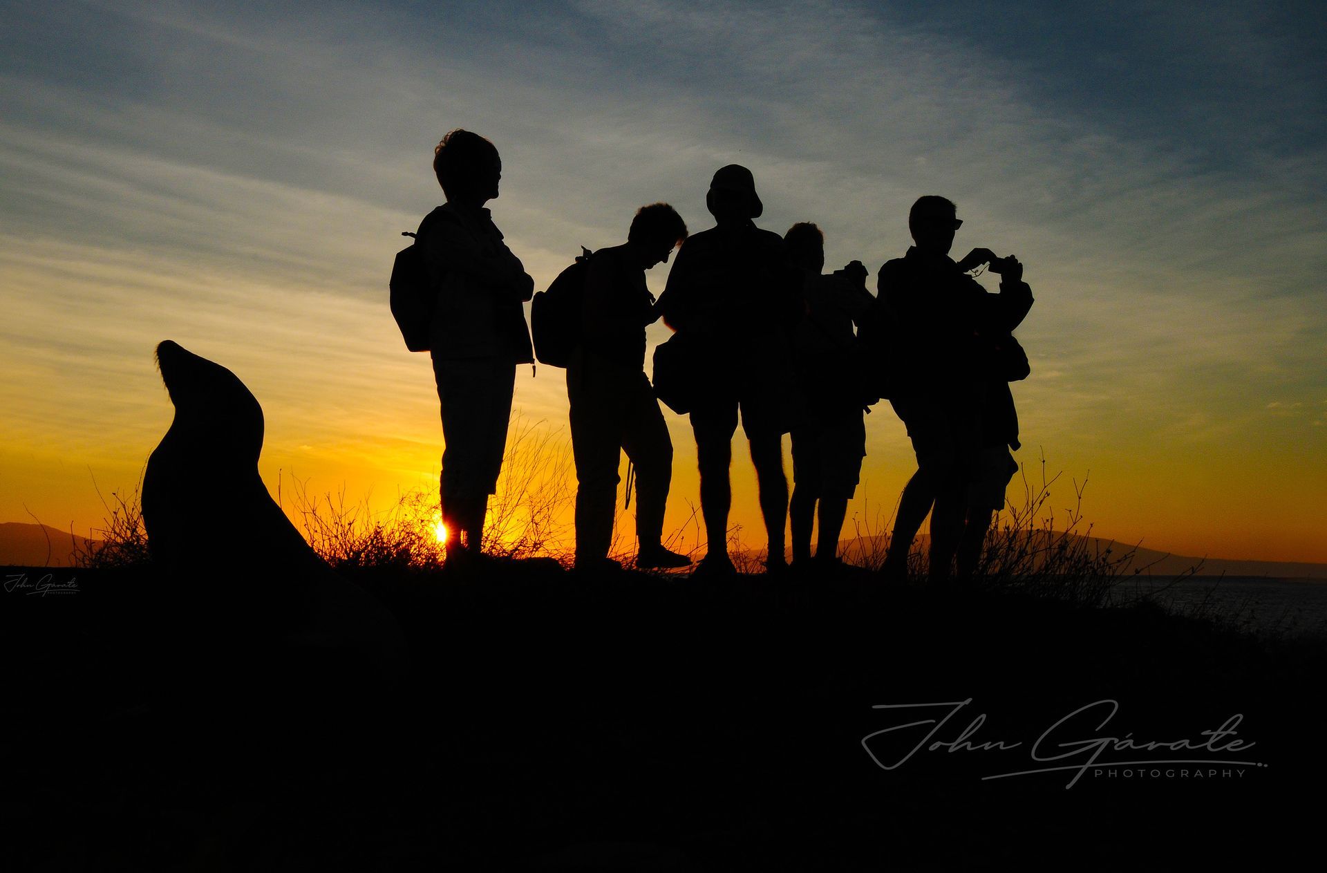Silhouetted group standing on a ridge at sunset, with orange sky and one seated figure at left