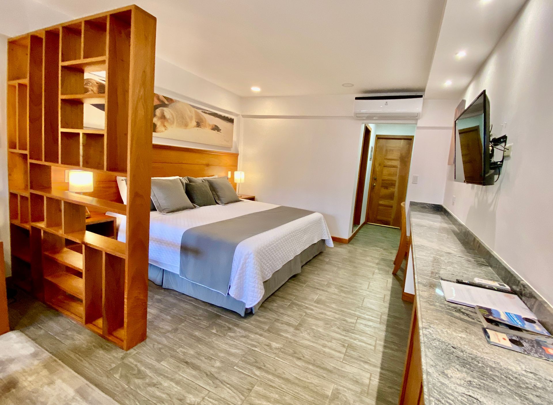 Modern hotel room with a bed, wooden shelving divider, long desk, wall TV, and hallway door in the Galapagos Islands.