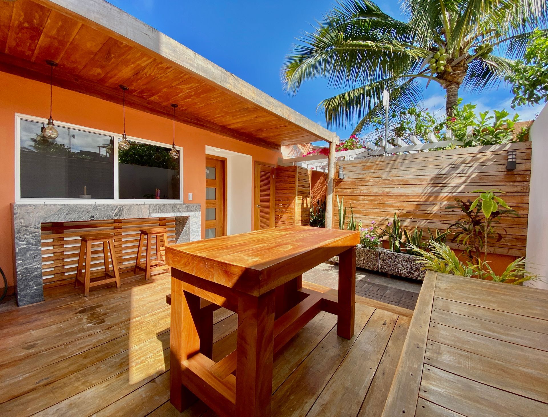 Sunny Galapagos hotel wooden patio with picnic table, bar seating, and tropical plants beside a modern house