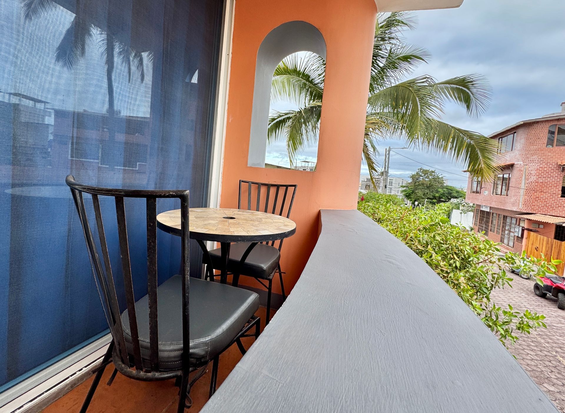 A Galapagos Hotel Balcony with two black chairs and a round table beside an orange wall, overlooking greenery and buildings
