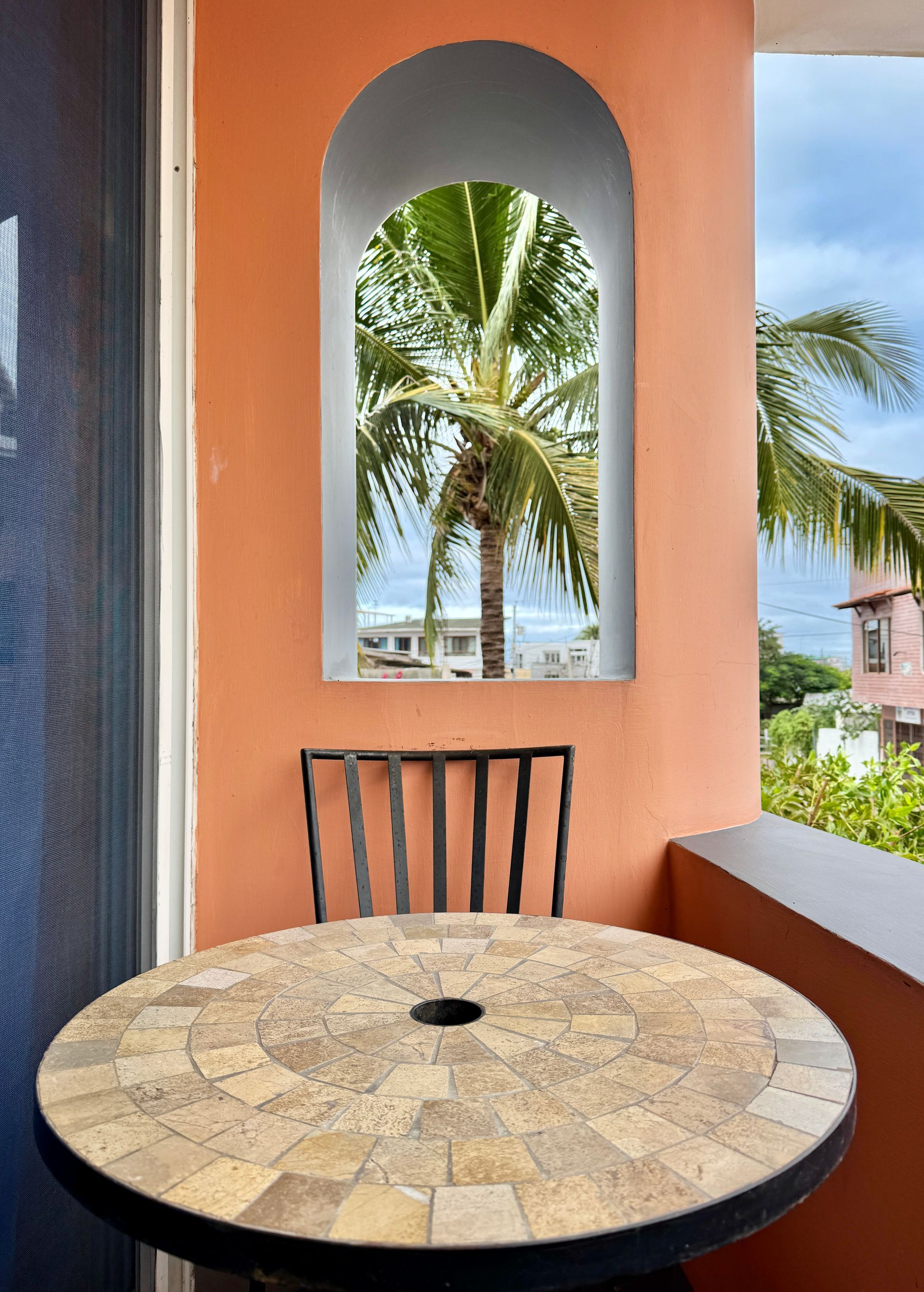 A Galapagos Hotel balcony with a mosaic-tiled table and a black metal chair. An arched window frames a palm tree and ocean view.