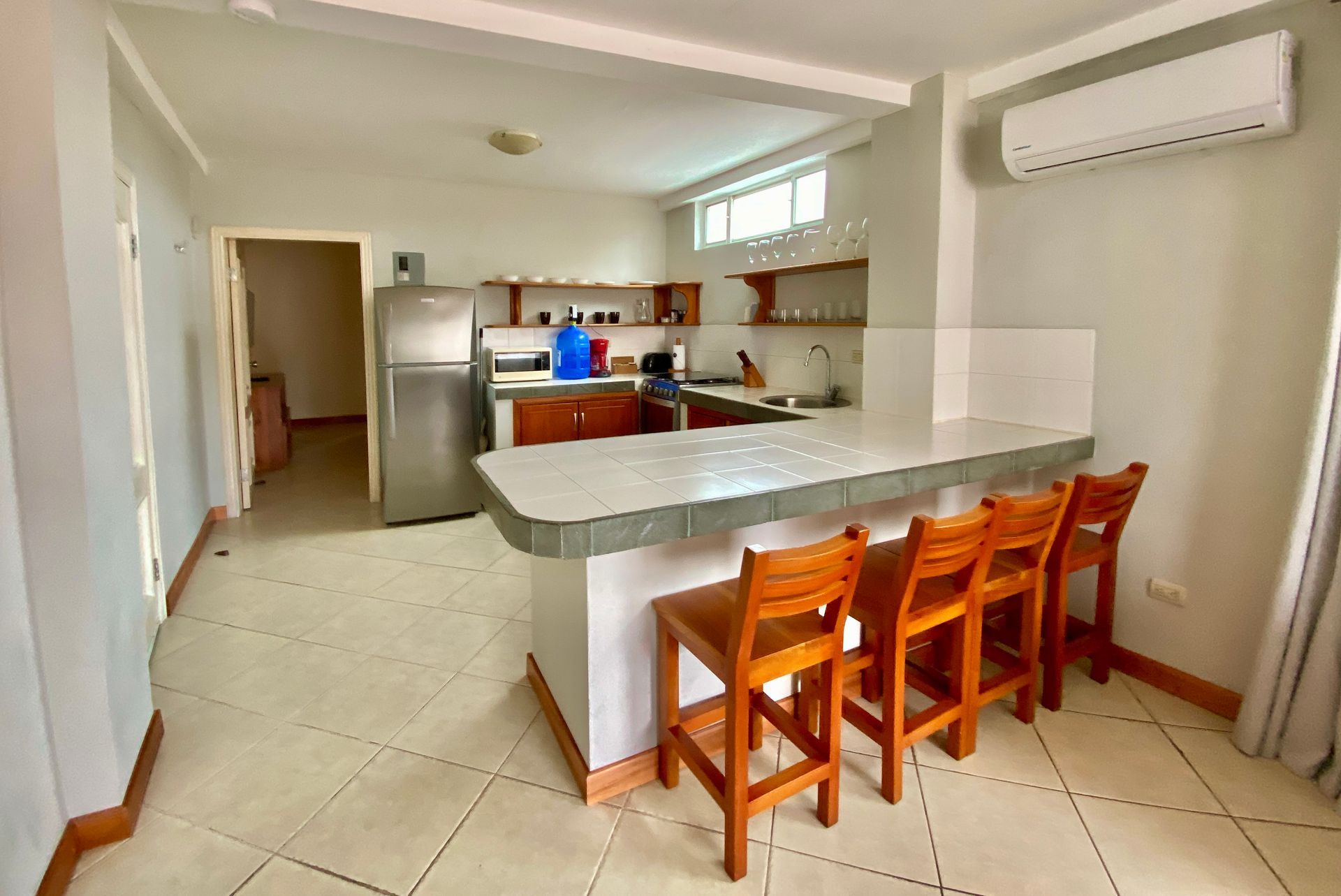 Bright hotel kitchen with a gray island, wooden stools, stainless fridge, and wall-mounted air conditioner in the Galapagos Islands.