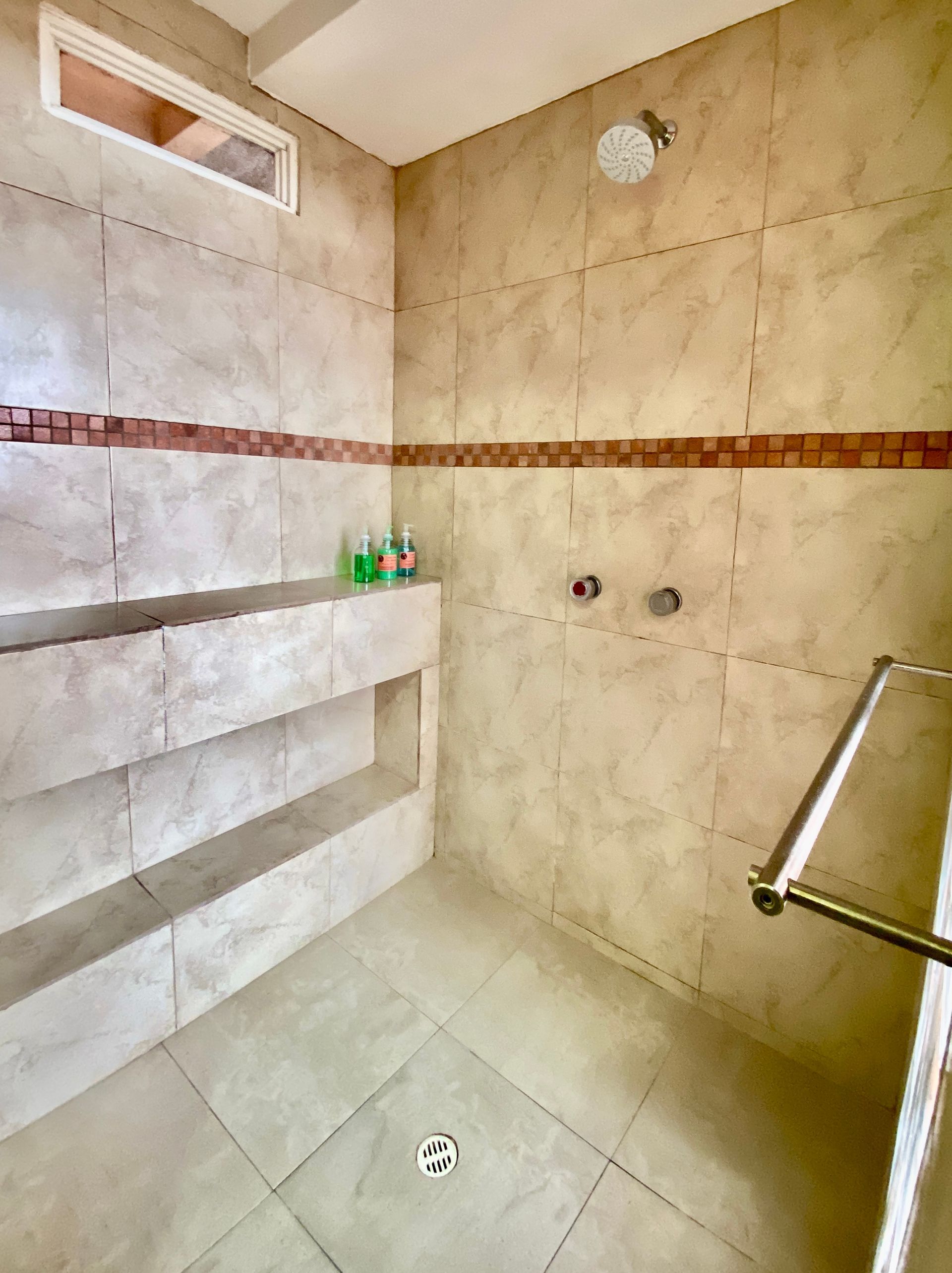 Tiled hotel shower stall with wall shelf, drain, and metal grab bar, in beige and white tones in the Galapagos Islands