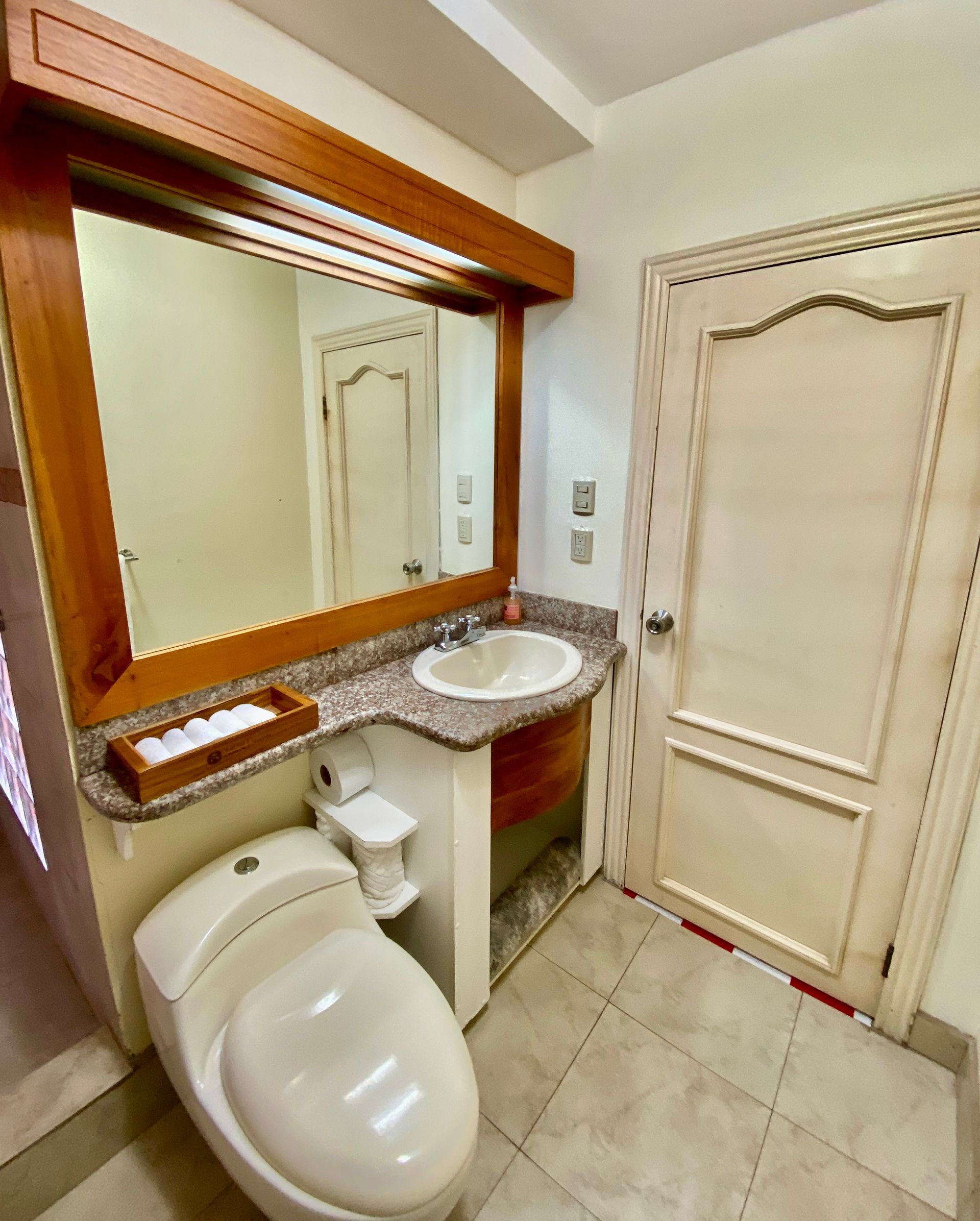 Galapagos Hotel bathroom with toilet, sink, large mirror, and closed door, in beige tile and wood trim.
