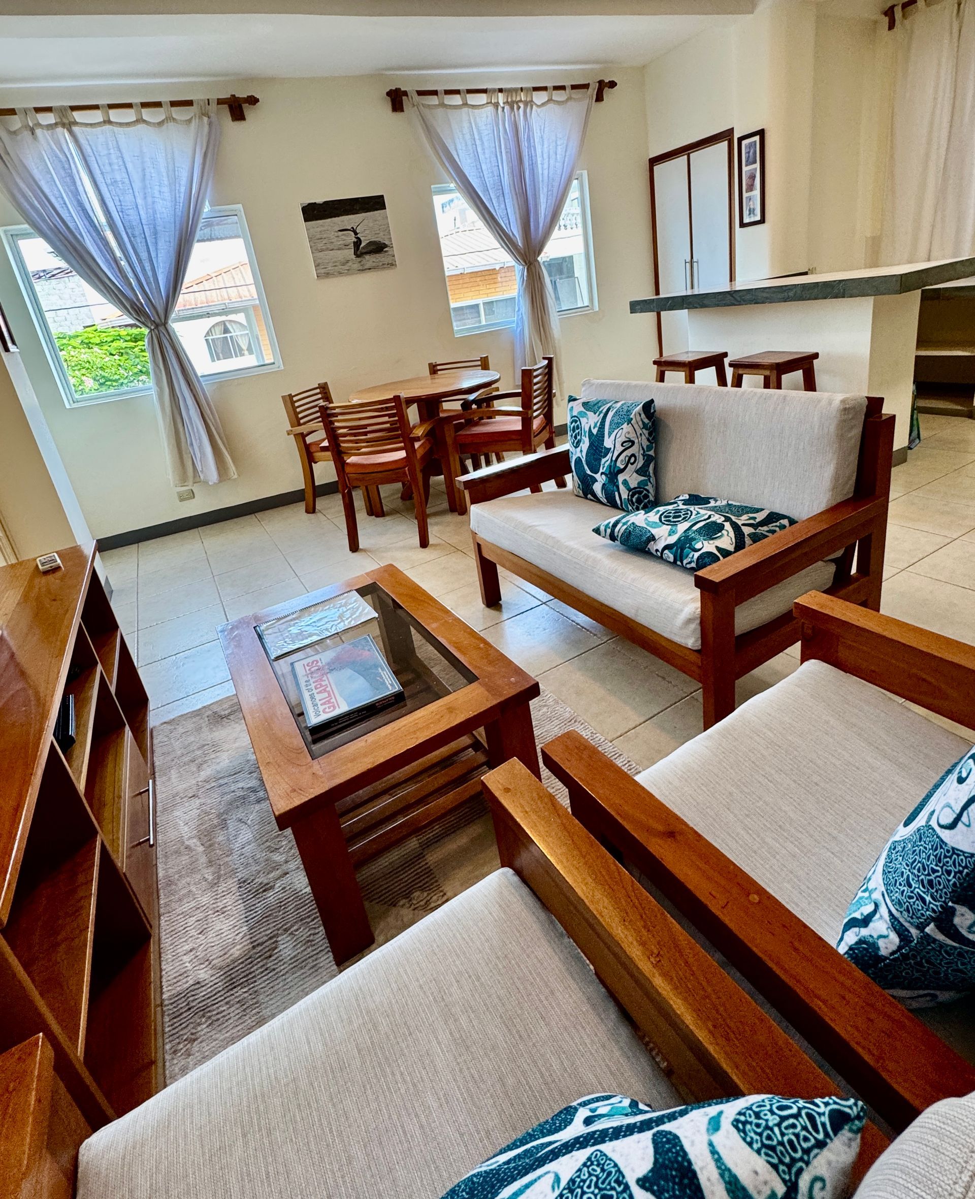 Bright Galapagos Hotel living room with wooden furniture, beige sofas, blue pillows, and a dining table by curtained windows