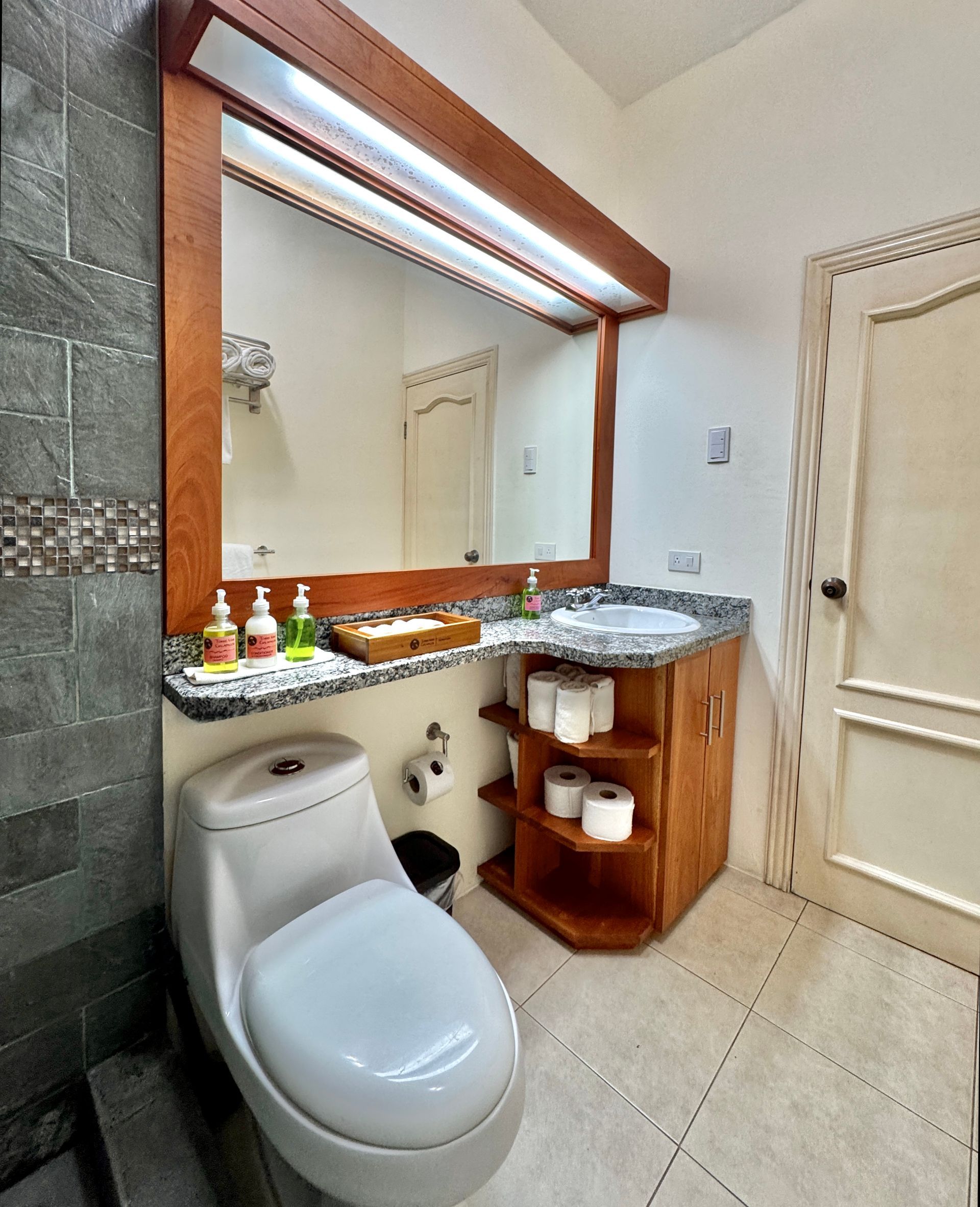A Galapagos Hotel bathroom with toilet, granite vanity, large mirror, beige tiled floor, and wooden shelving.