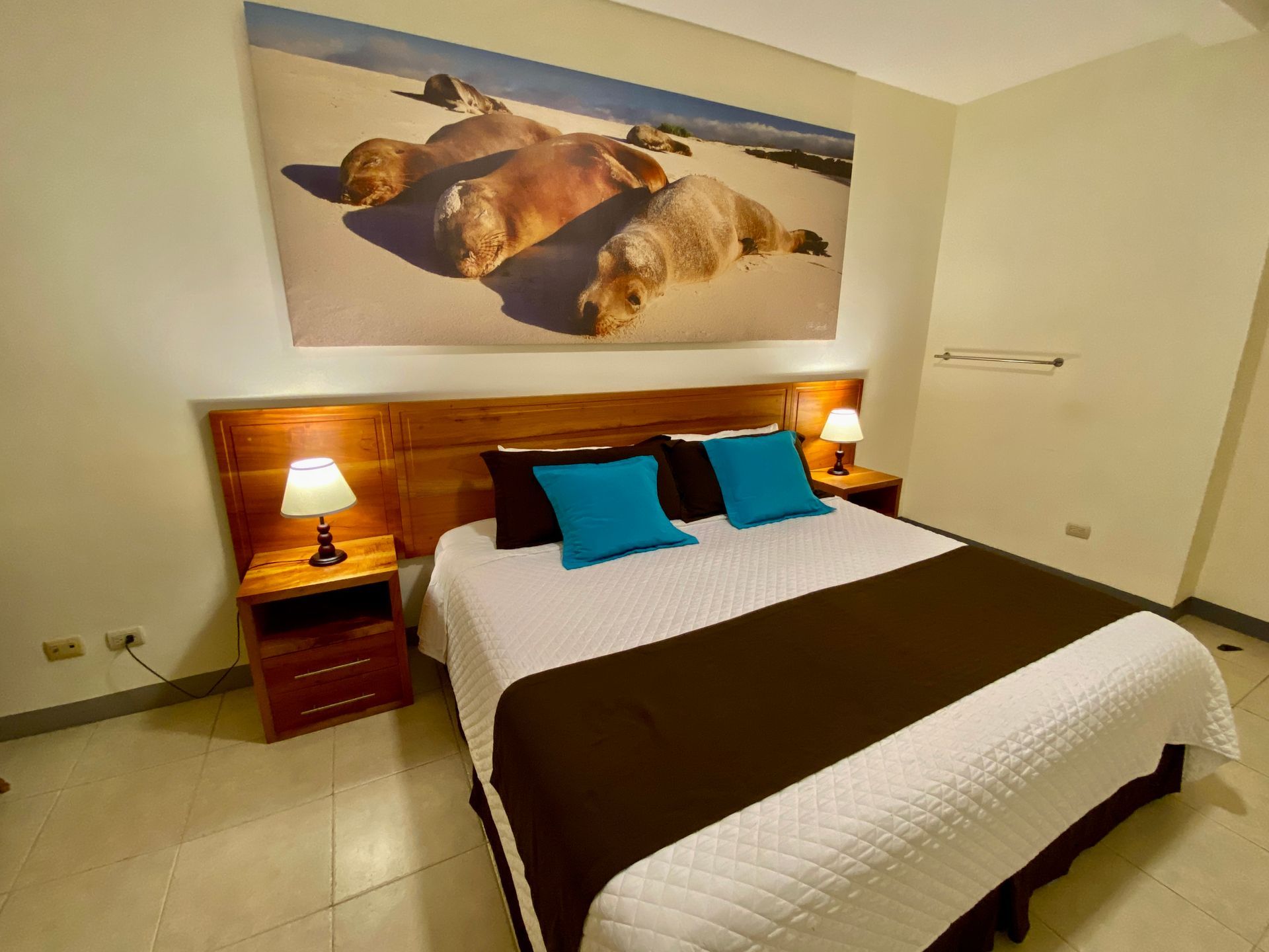 A Galapagos Hotel room with double bed, blue pillows, brown runner, and bedside lamps beneath wall art