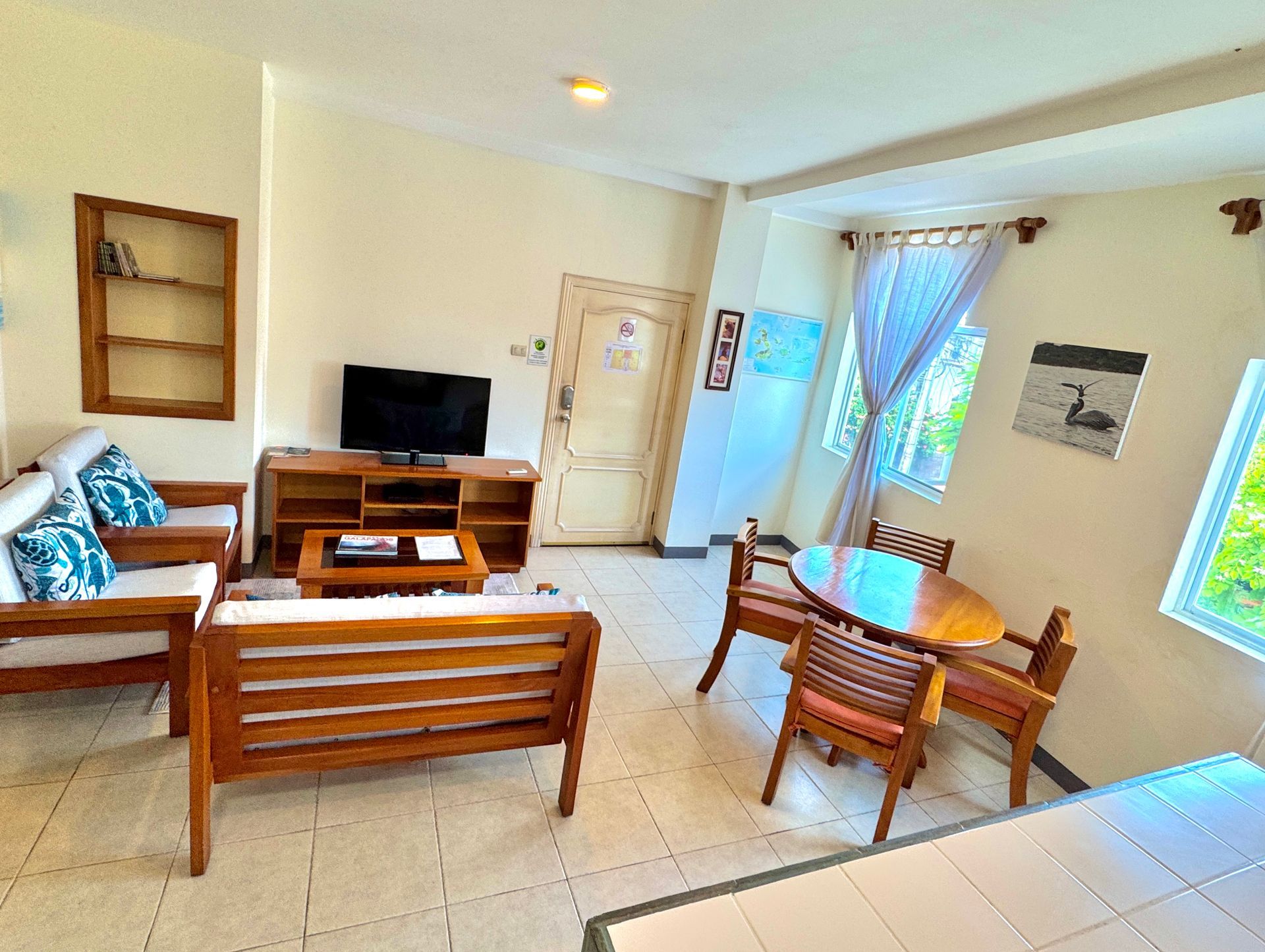 A Galapagos Hotel Bright living room with sofa, wooden TV stand, chairs, table, and sunlit windows.