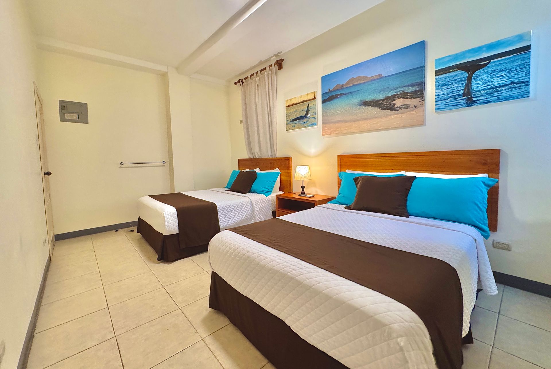A Galapagos Hotel room with Two double beds with brown and white bedding in a well-lit room with beach landscape art on the wall.