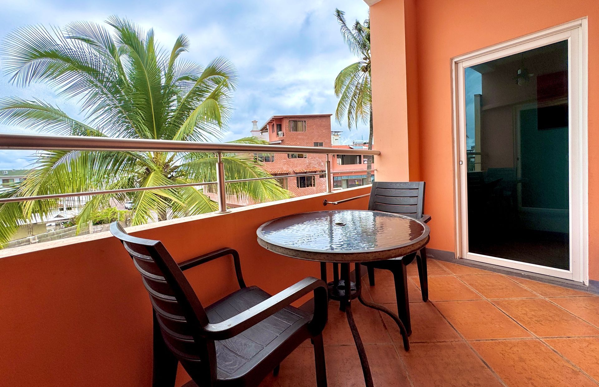 A Galapagos hotel Balcony with round table, two chairs, orange walls, and palm trees beyond the railing.