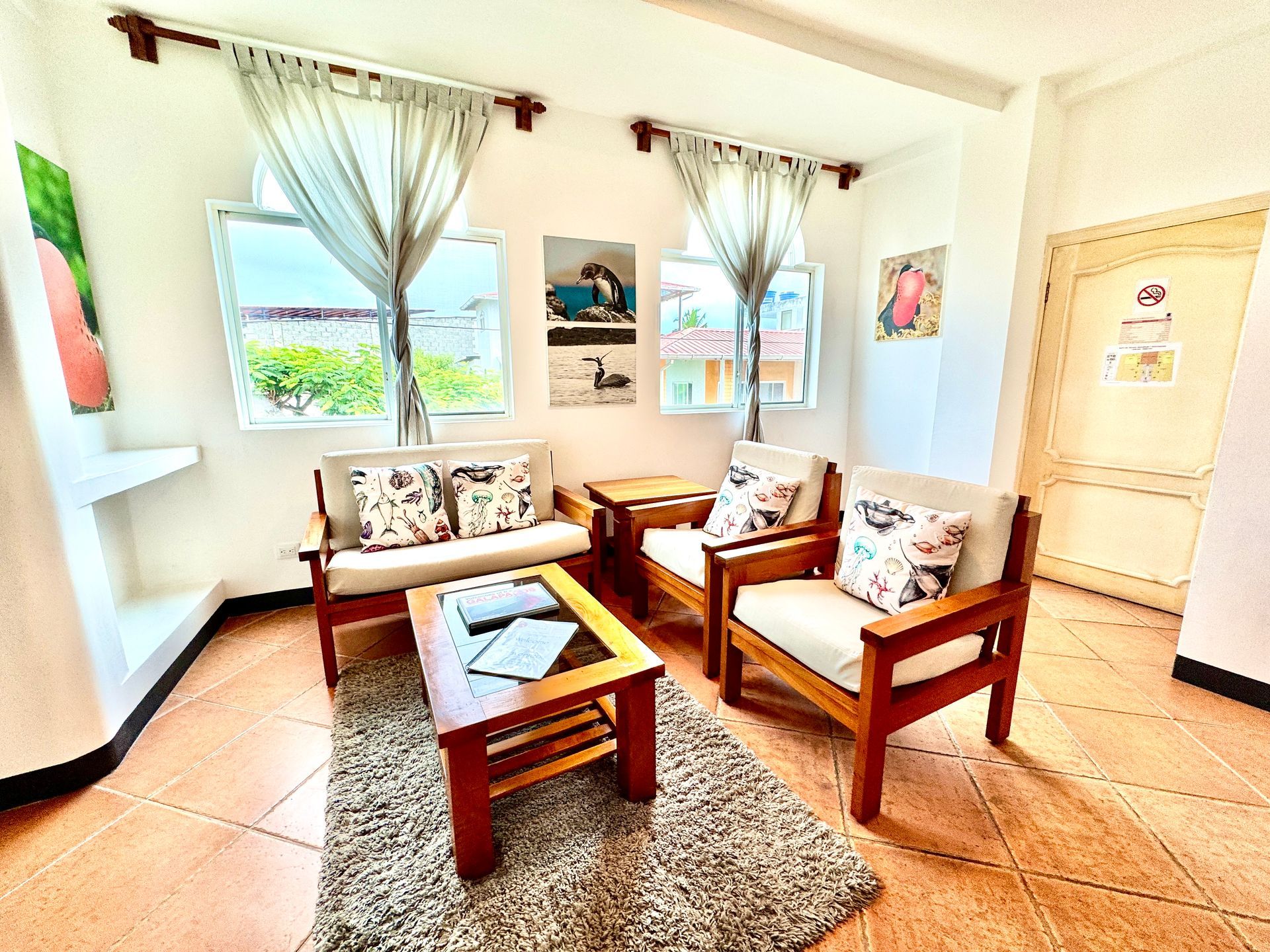 A Galapagos hotel living room with wooden furniture, cream cushions, and a small rug. Windows provide natural light.
