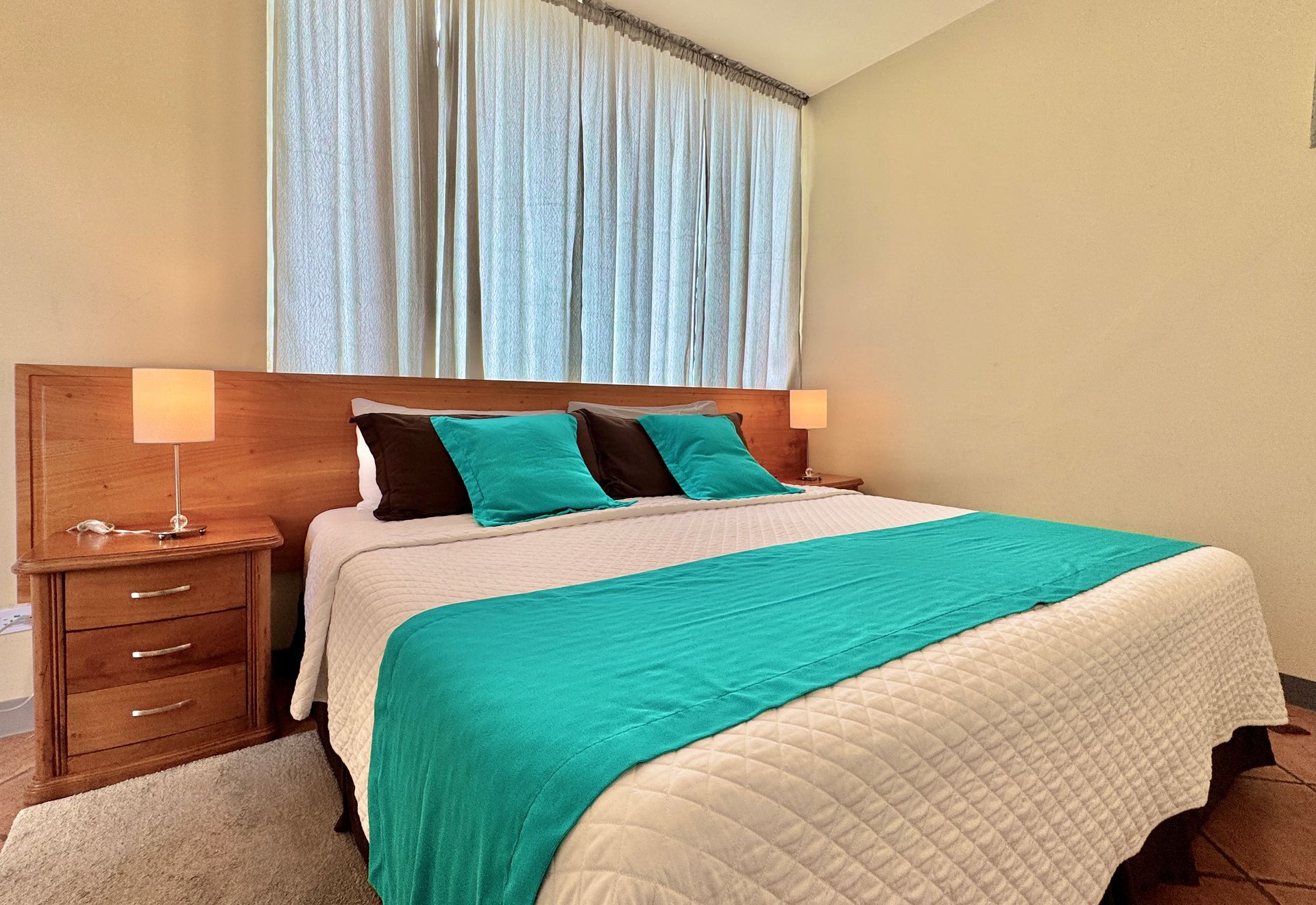 Galapagos hotel Bedroom with a large bed, teal accents, two nightstands with lamps, and a window with curtains.
