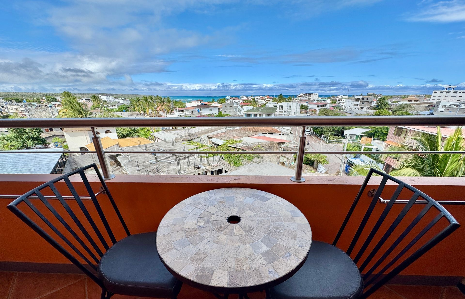 A Galapagos hotel balcony with a round table and chairs overlooking a city and the ocean under a blue sky.