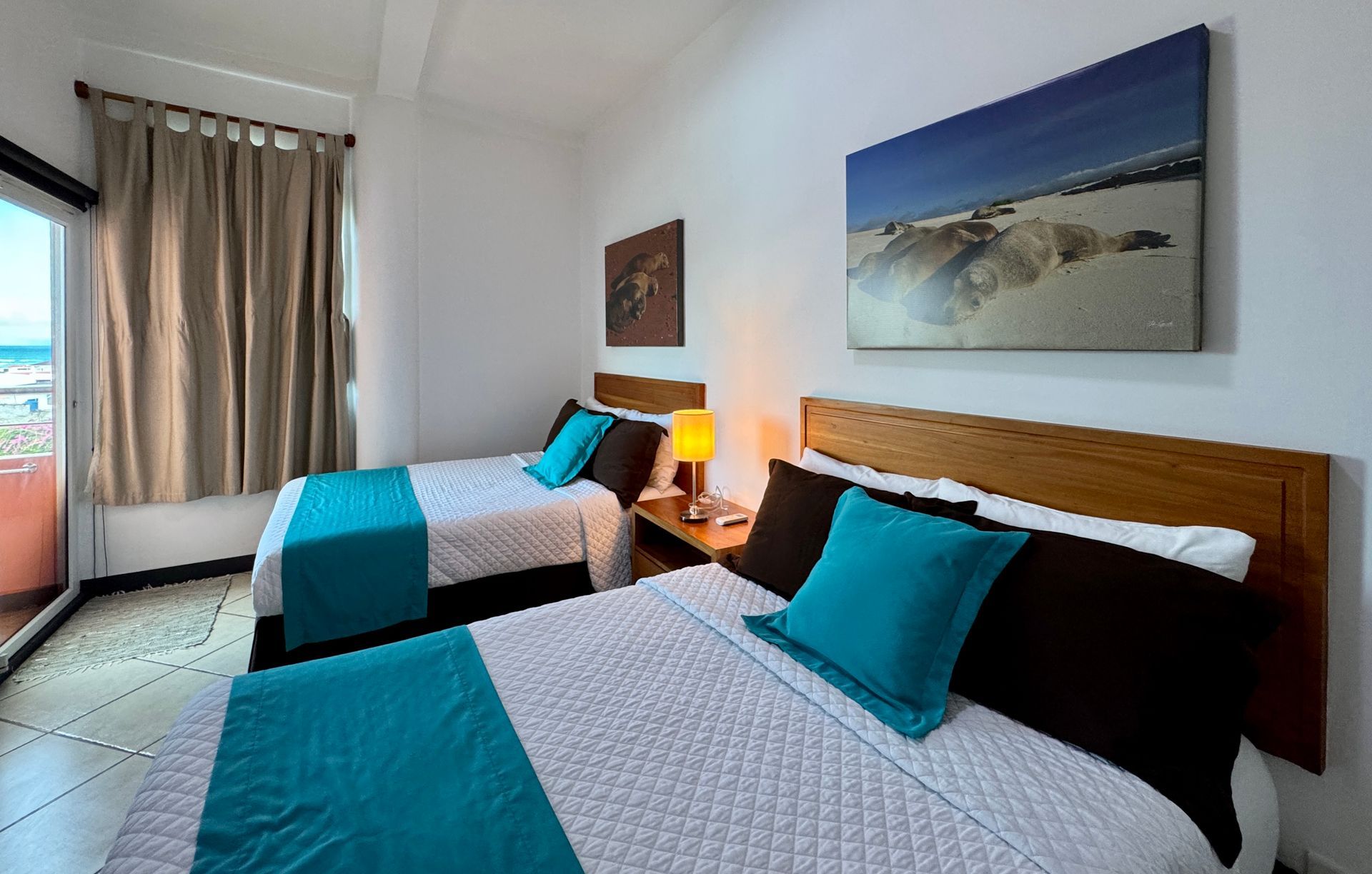 Two double beds in a bright Galapagos hotel bedroom. Teal blankets and pillows contrast with the white bedspreads