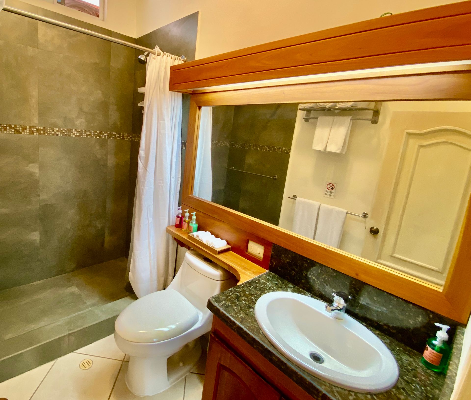 A Galapagos Hotel Bathroom with a toilet, sink, shower, and large mirror. Wooden accents, grey tile, and white towels.