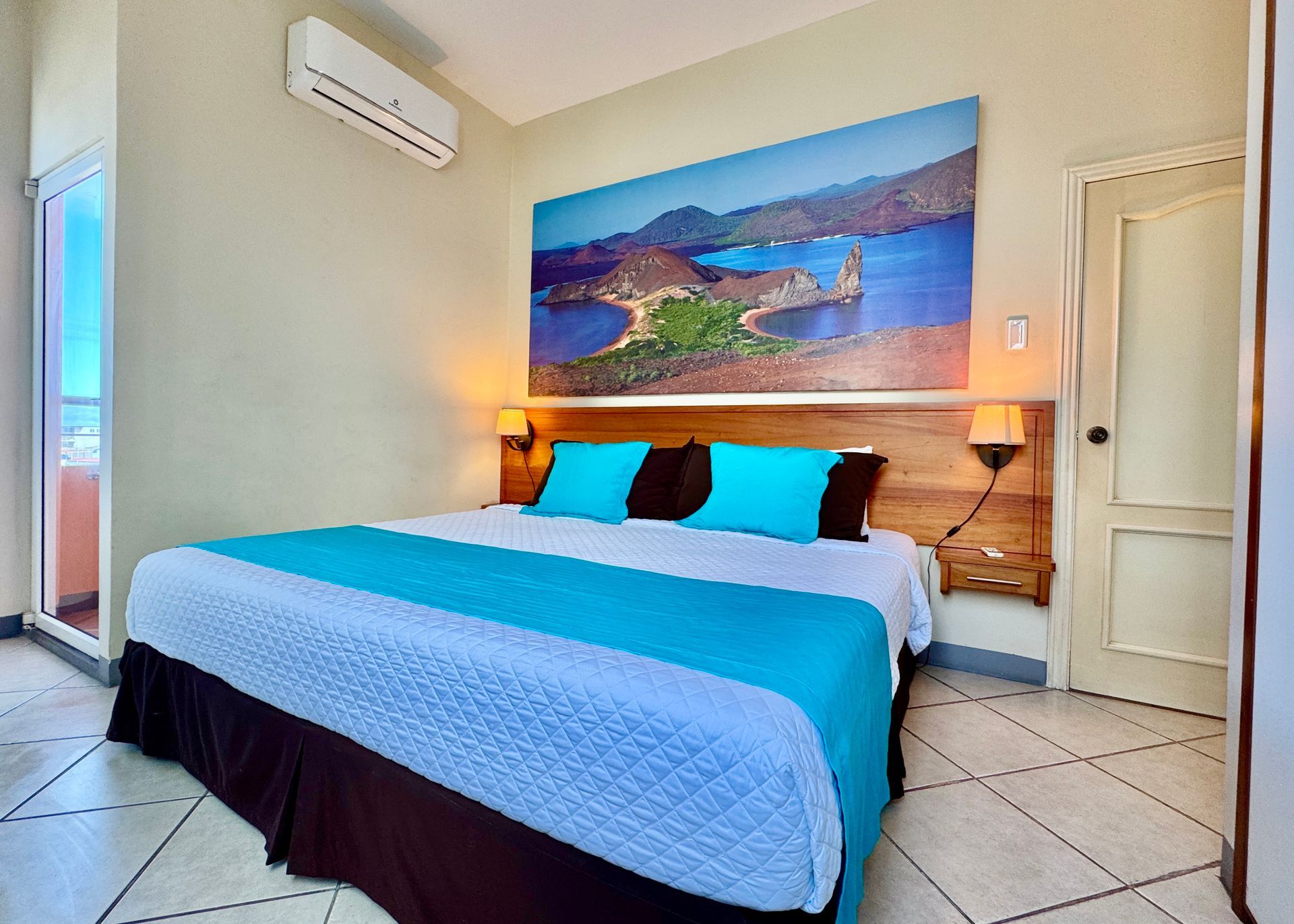 A Galapagos Hotel bedroom with a large bed, turquoise bedding, and a landscape photo above the headboard.