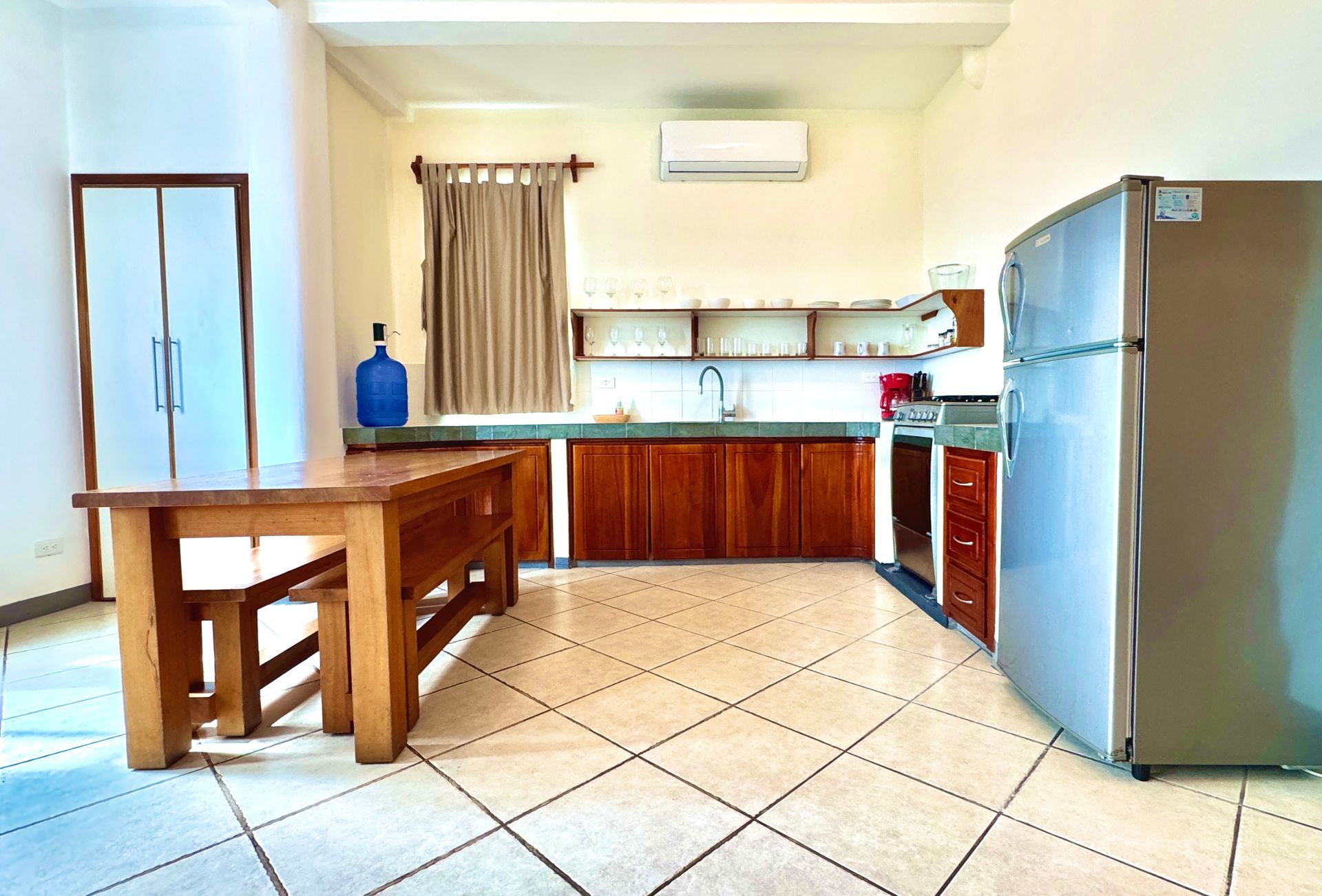 Bright Galapagos hotel kitchen with wooden table, tiled floor, cabinets, sink, and silver refrigerator.