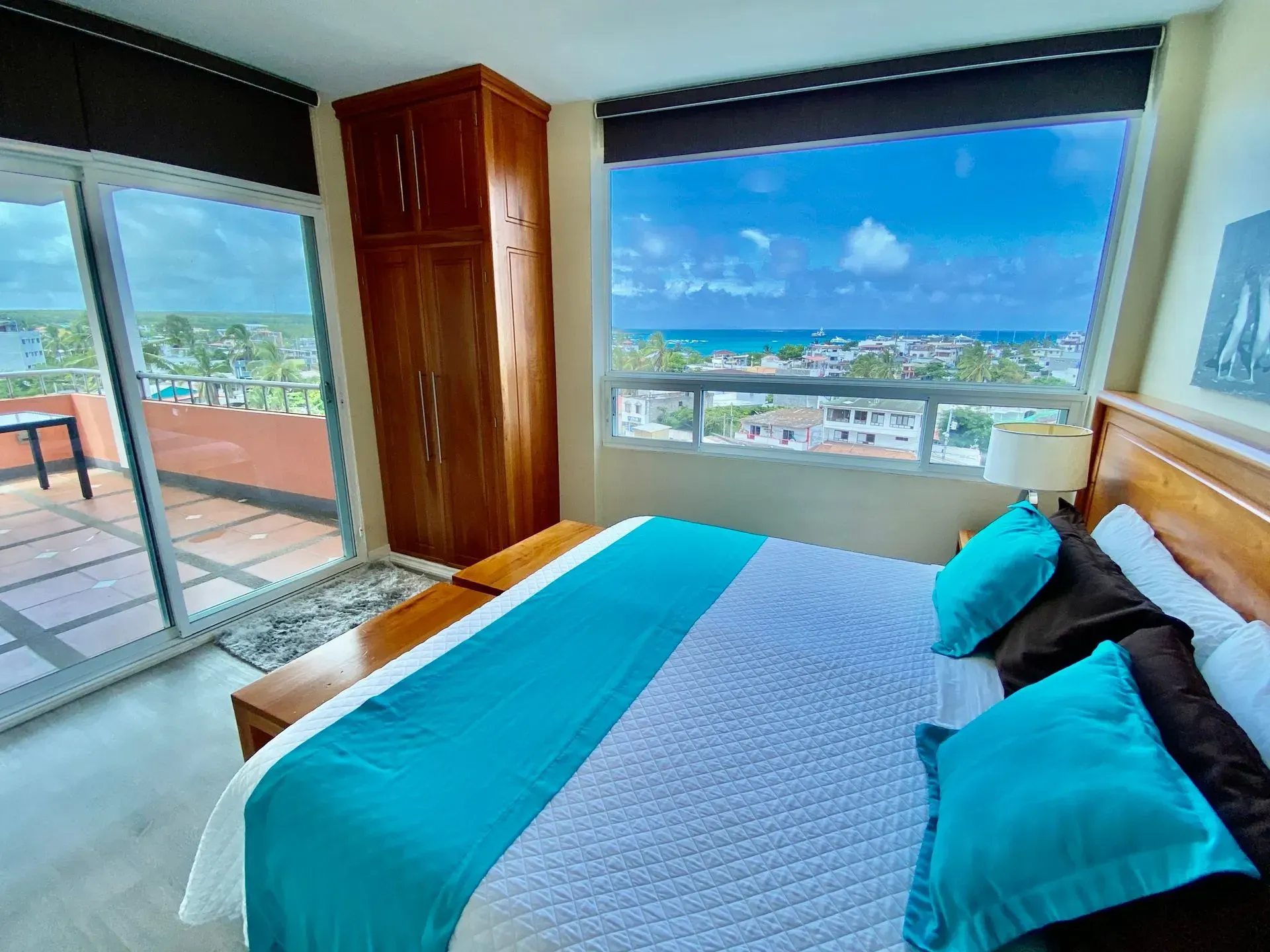 Bright bedroom with turquoise bedding, large windows, and ocean-view balcony under a blue sky