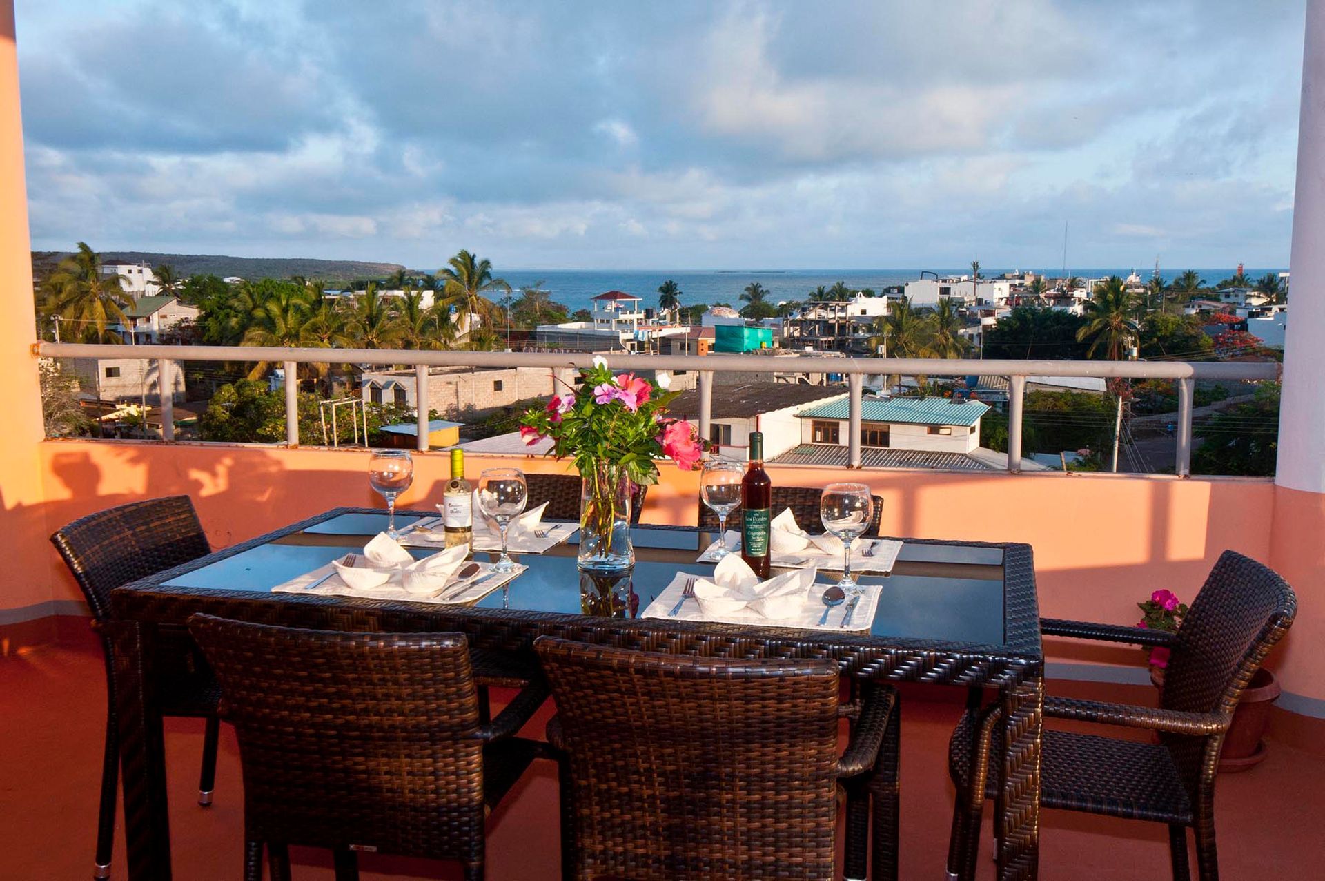 A Galapagos hotel Rooftop dining table with flowers and drinks, overlooking a coastal town and ocean at sunset.
