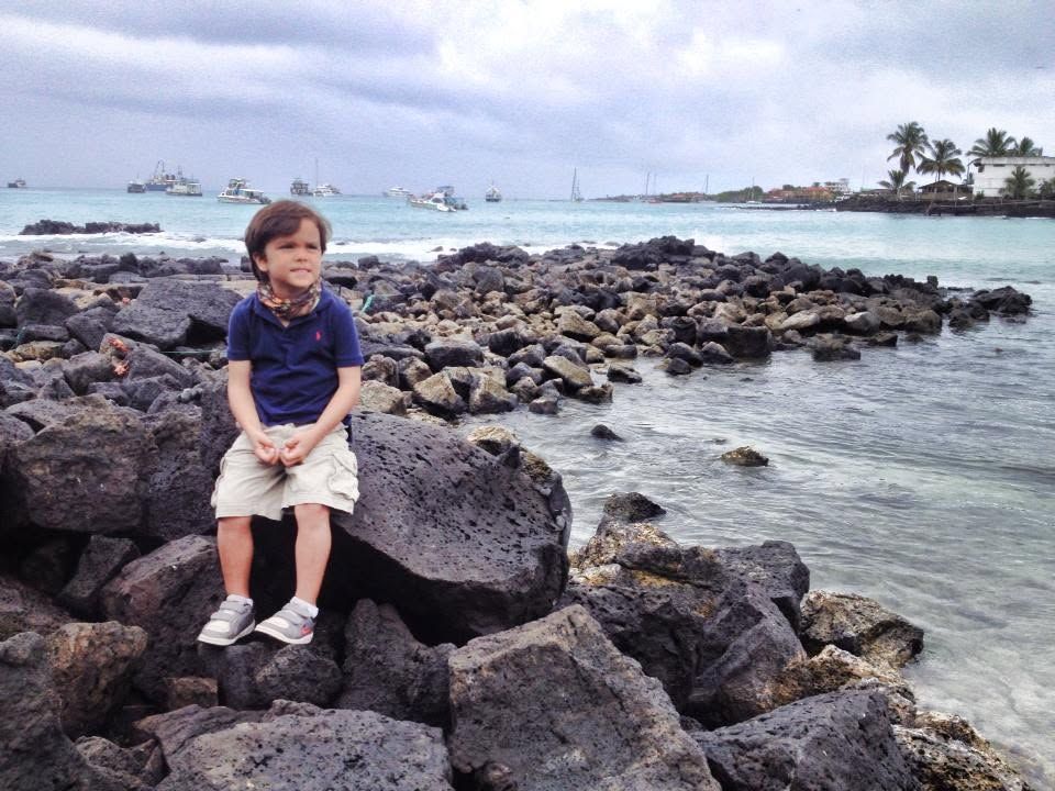 Child sitting on dark rocks by the ocean, with waves and boats in the background.