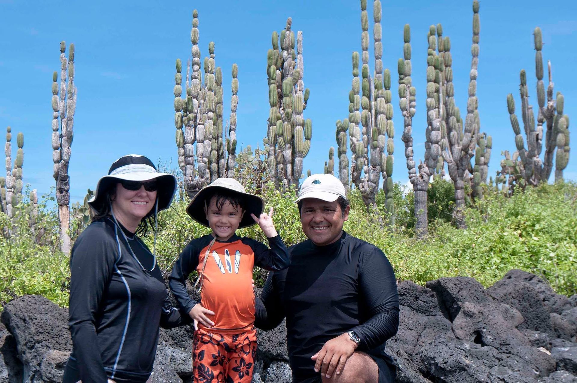 Three people posing on black lava rocks with green shrubs and tall cactus-like plants under a bright blue sky.