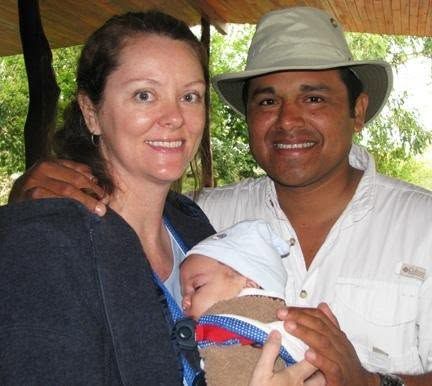 Two adults smiling and holding a sleeping baby outdoors under a canopy