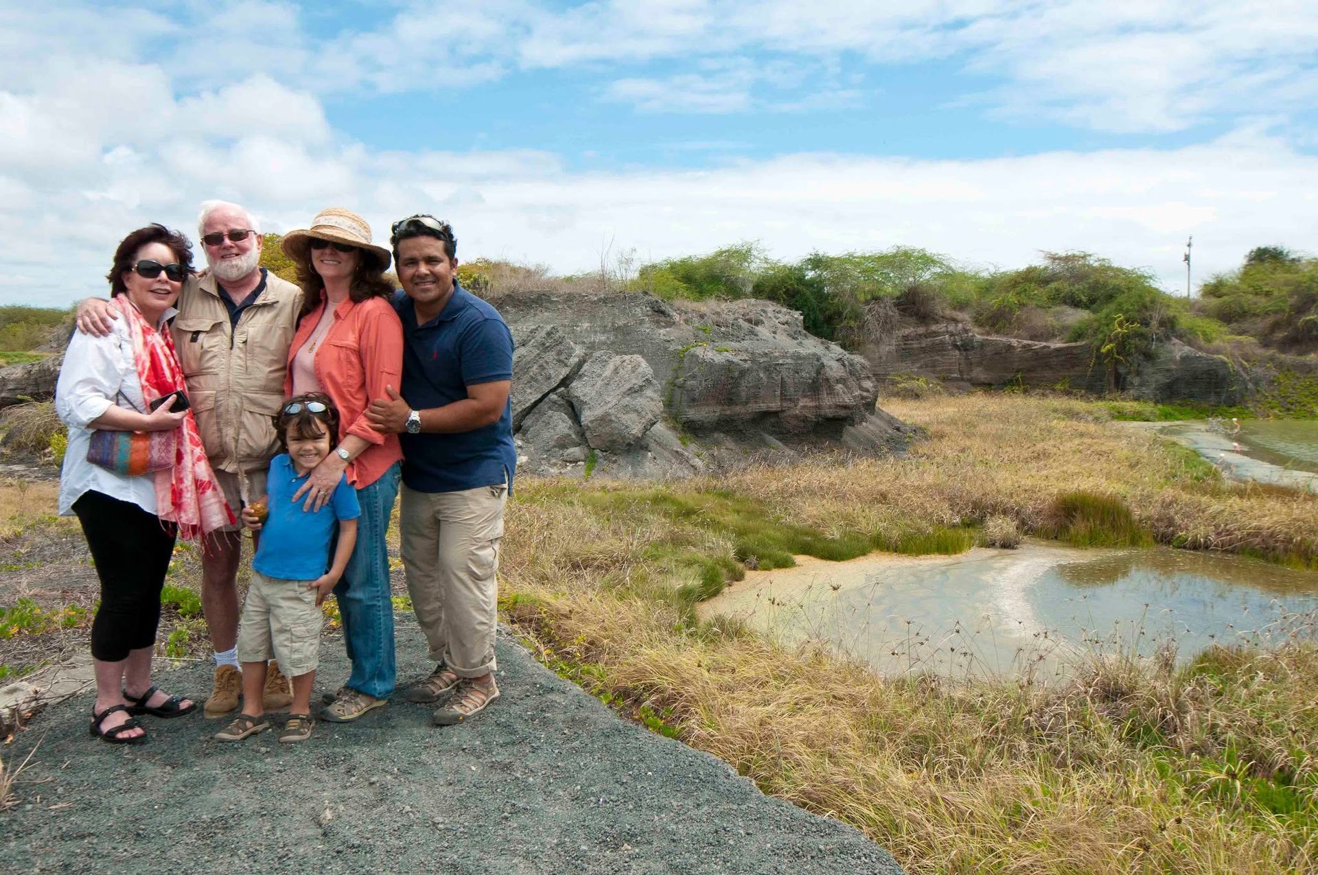 Group of four posing by a rocky pond on a dry, scrubby hillside under a bright sky