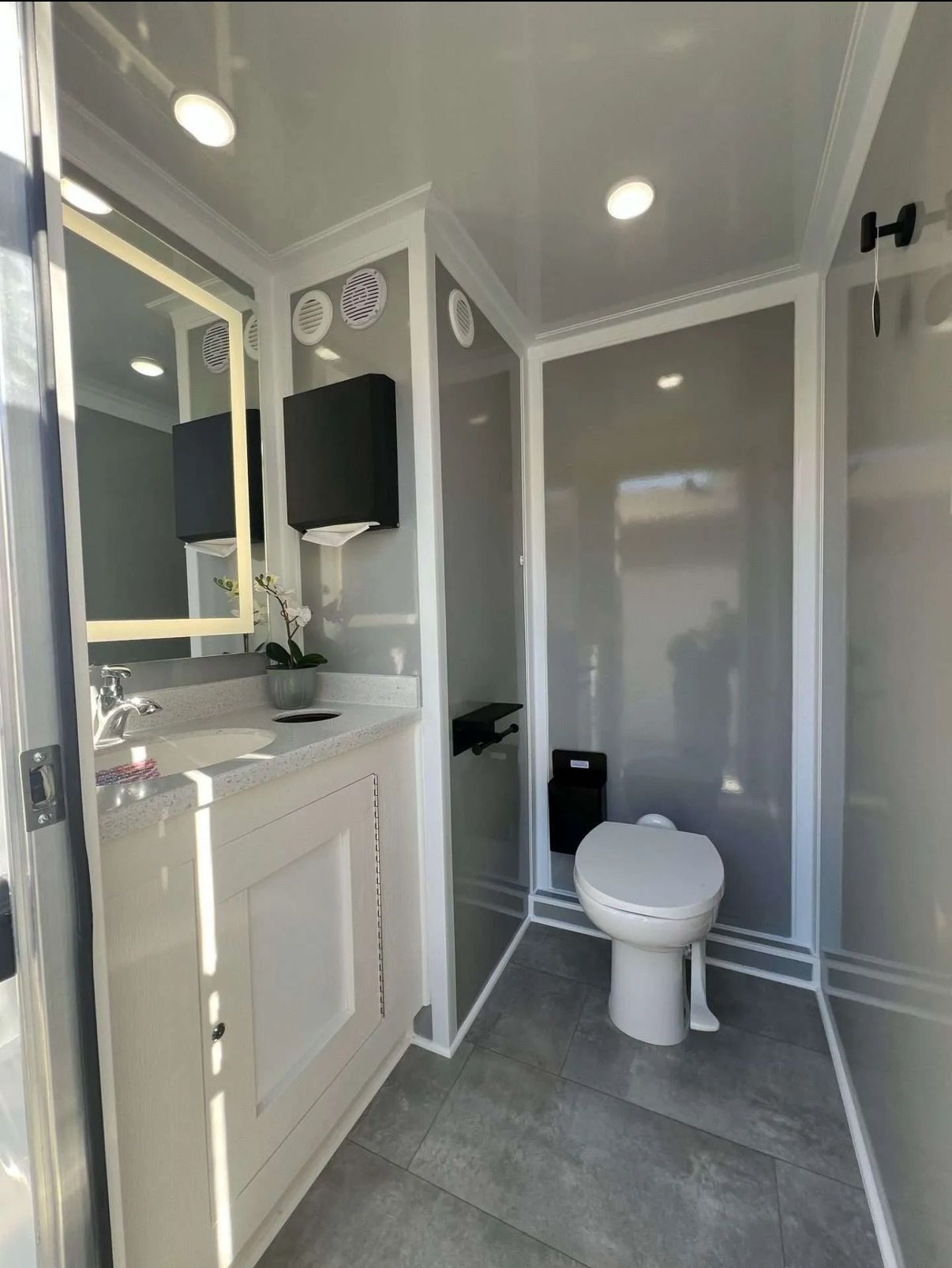 Interior of a luxury portable restroom with a sink, toilet, mirror, and modern gray and white decor.